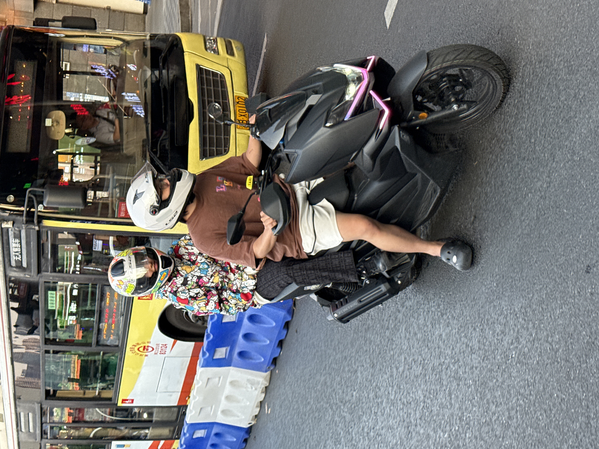 Two people on a black Zontes motorcycle are stopped on a street, presumably in Yuzhong District, China. The driver wears a brown shirt, white shorts, a white helmet, and black Crocs. The passenger wears a helmet and a colorful, patterned jacket. A yellow bus with Chinese characters and number 83124 is directly to their left. The bus has a visible driver and several passengers. Blue and white plastic barriers are positioned in front of the bus. The street is gray asphalt. The motorcycle has pink and black accents.