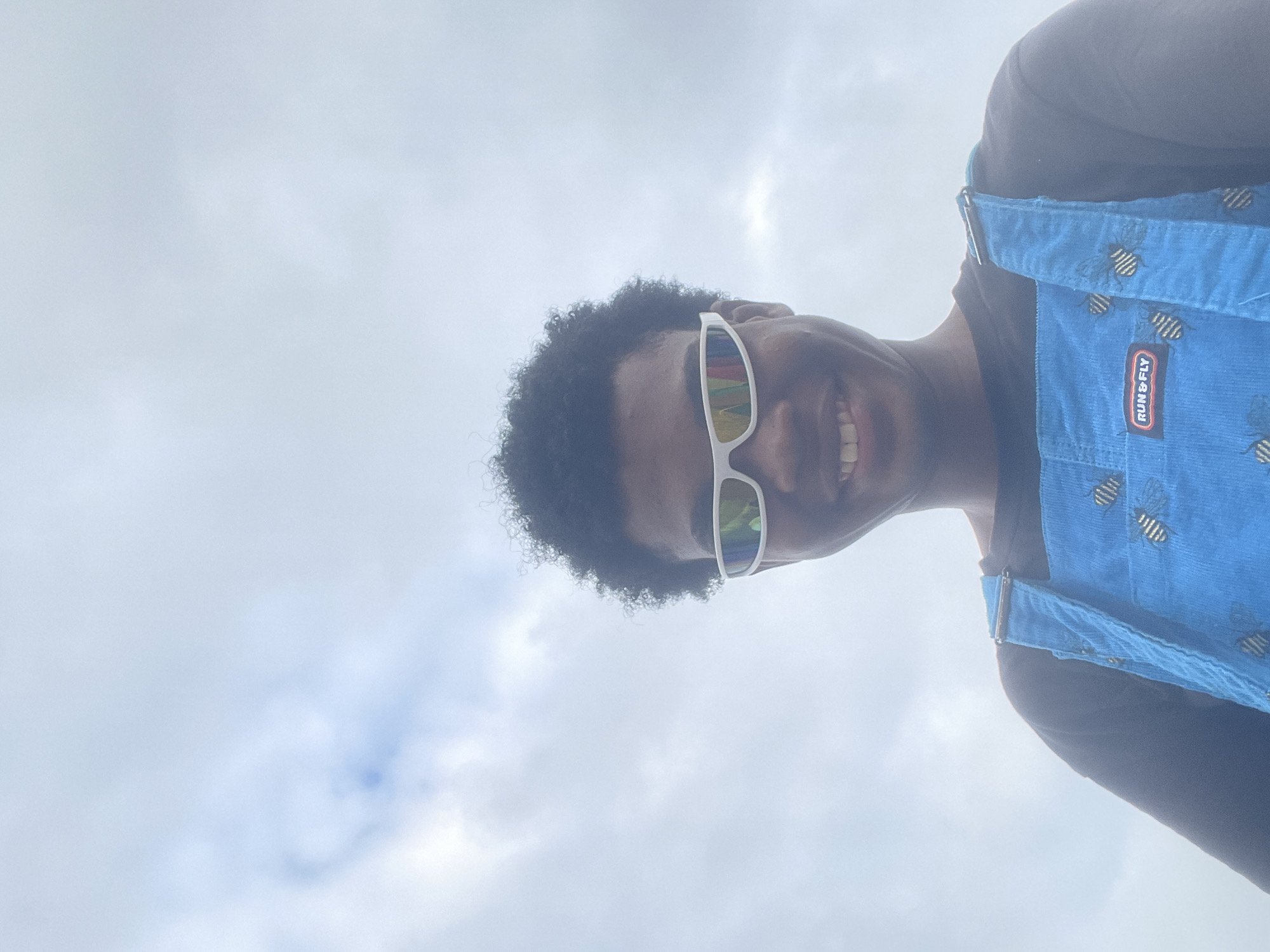 An individual with dark, curly hair is taking a selfie outdoors against a cloudy sky. They are wearing white, wraparound sunglasses with a rainbow-colored tint and a blue corduroy overall with a pattern of bees. A black t-shirt is visible underneath the overalls, and on the bib of the overalls, there is a red and white tag that reads "RUN & FLY." The individual appears to be smiling. The location is Inupa Kekere, Nigeria.