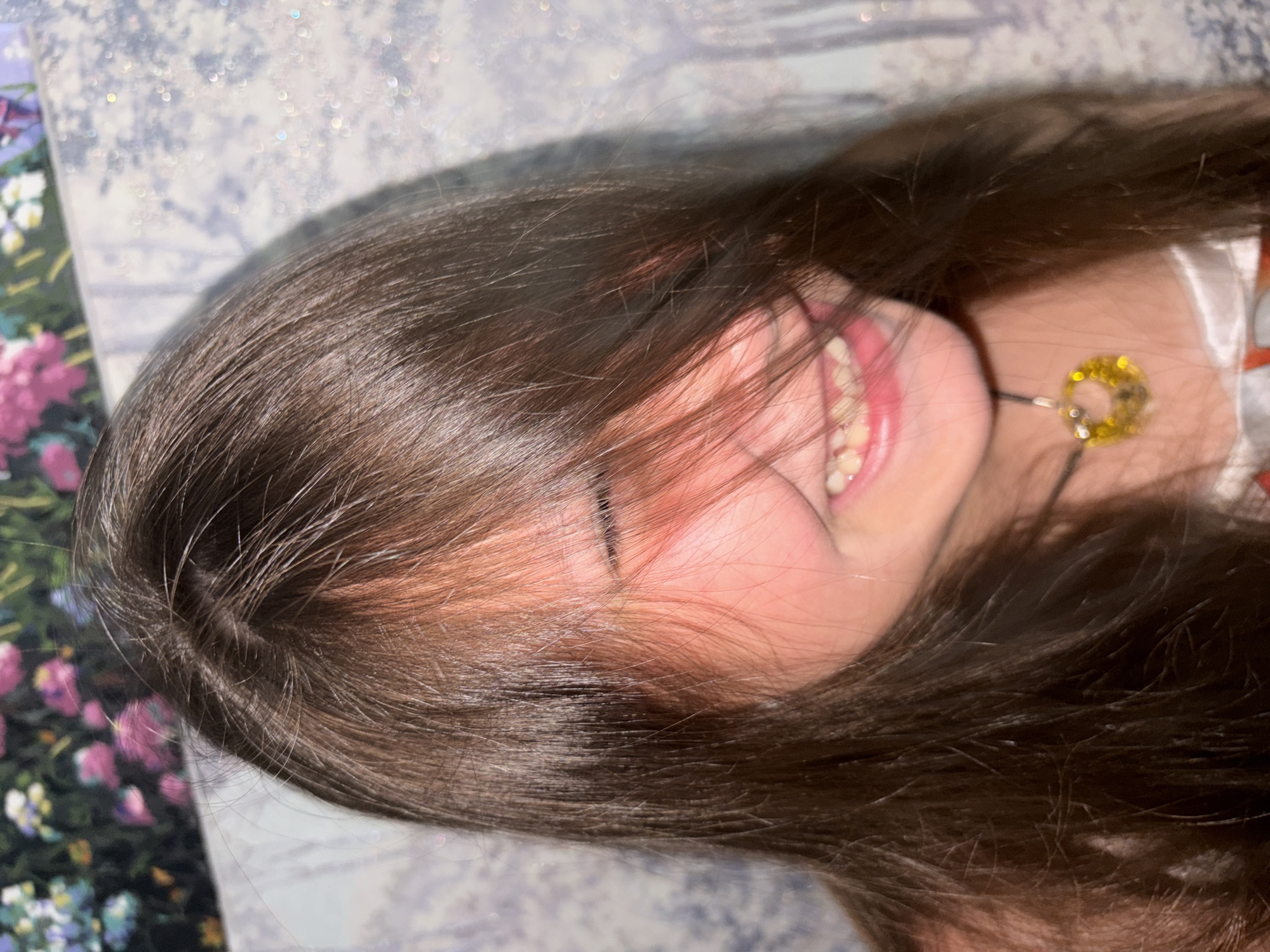 A close-up, rotated view of a young person with long brown hair partially obscuring their face. The person is smiling, revealing their upper teeth. They are wearing a necklace with a circular pendant that appears to be gold. The background is a patterned surface that looks like fabric or bedding. The location is not discernible from the image.