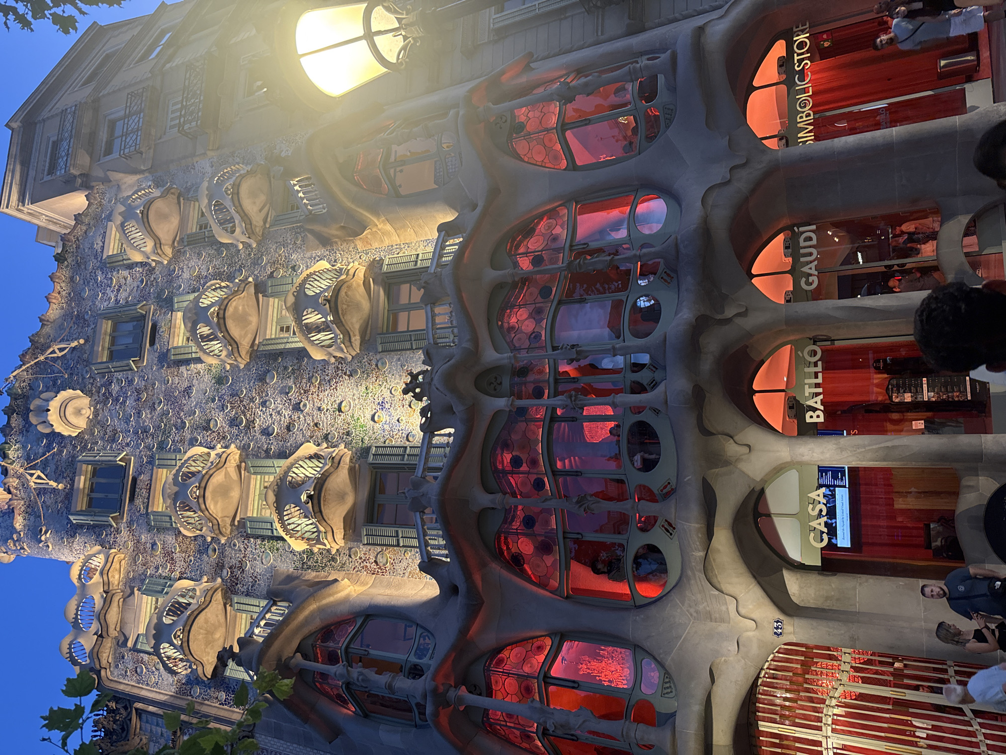 This is a low-angle, exterior, nighttime shot of Casa Batlló, located on Passeig de Gràcia in Barcelona, Spain. The building's facade, designed by Antoni Gaudí, is illuminated, highlighting its characteristic undulating organic forms and colorful mosaic tiles. Several large, arched windows on the lower levels glow with a vibrant red light, revealing intricate stained-glass patterns. Above these, a series of balconies protrude, each with a unique, bone-like structure, resembling masks or skulls. The upper floors feature smaller windows with green shutters and more of the textured, mosaic-covered walls. A street lamp with a warm yellow glow is visible on the right side of the frame, partially obscuring the building. At the base of the structure, signage for "CASA BATLLÓ" and "GAUDÍ" are visible above entrances draped with red curtains. Several people are visible in the foreground and near the entrances, dressed in casual attire, appearing to be tourists or passersby. The sky above is a deep blue, indicating dusk or early evening. The architecture is the primary subject, with the people serving as contextual elements.