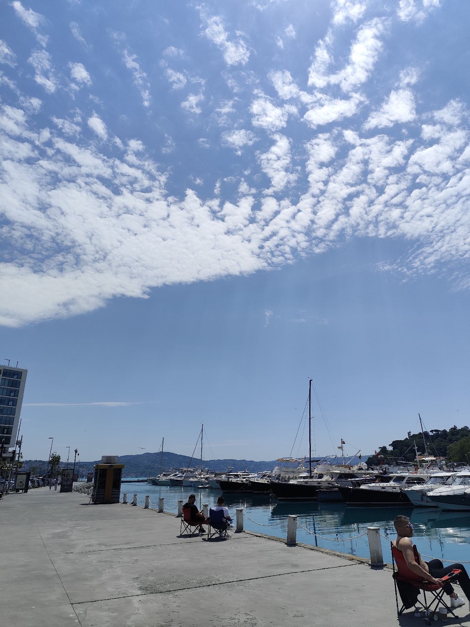 The image portrays a bright and sunny day at a vibrant marina, characteristic of a coastal setting like Sarıyer, Turkey. The foreground features a broad, light-colored concrete pier or promenade extending into the midground. The sky above is a clear, brilliant blue, dotted with numerous small, fluffy white clouds, indicating pleasant, fair weather and suggesting it is midday.
Three men are visible on the pier, enjoying the waterfront. In the midground-left, two men are seated in foldable chairs—one red, one blue—facing the tranquil, turquoise water of the marina. They appear to be relaxing, possibly observing the activity or fishing, though no fishing equipment is clearly visible. Closer to the foreground on the right, a third man, wearing a red tank top and sunglasses, sits in another red foldable chair, also facing the water, seemingly unwinding and soaking in the sun.
The marina is bustling with a variety of vessels, predominantly motor yachts and some sailboats, neatly docked along the pier. Their masts and structures rise against the backdrop of distant green hills across the water. The edge of the pier is marked by white posts interconnected with chains. On the far left, a modern building with numerous windows is partially visible, hinting at an urbanized waterfront area. A small, yellow and brown kiosk-like structure is also situated on the pier further down. Small birds can be seen flying gracefully over the water and in the sky.
There is no discernible text clearly visible on any signs or objects within the image. The overall impression is one of leisure, tranquility, and enjoyment of the scenic outdoor environment.