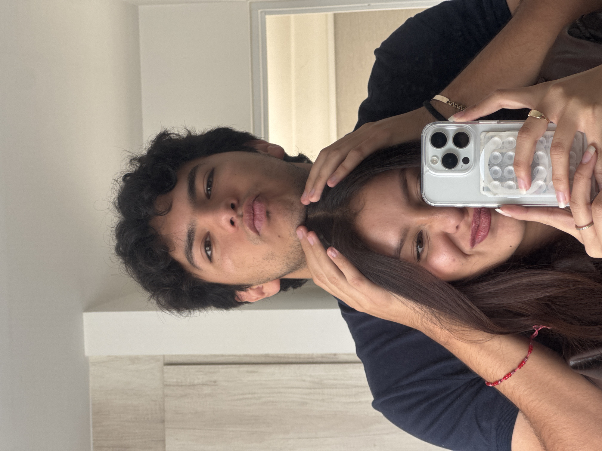 A young man and woman are posing for a selfie. The man has dark, curly hair and is puckering his lips. The woman has long, dark hair and is holding a phone with a clear case and grip stickers, her hand is adorned with rings and bracelets. They are both wearing dark-colored shirts. The location is not identifiable as San Francisco Ocotlán (Ocotlán), Mexico from the available information. The background includes a white wall and door.