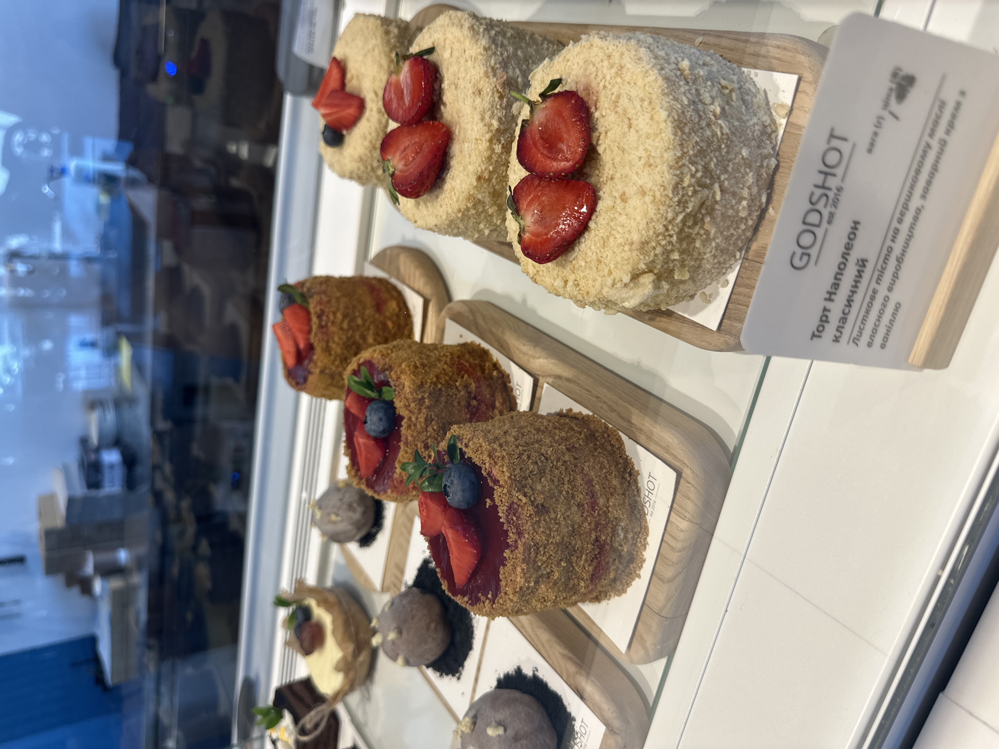 A display case in a bakery holds an assortment of round cakes topped with strawberries and blueberries, and some smaller pastries. The cakes are cylindrical with a crumbly exterior. One cake is topped with red jelly and berries, while others have a cream-colored coating and are decorated with sliced strawberries and blueberries. In the foreground, a white card with Ukrainian text, including "GODSHOT", describes one of the cakes. The background shows reflections of shelves and stacked items in a blurry, urban environment.