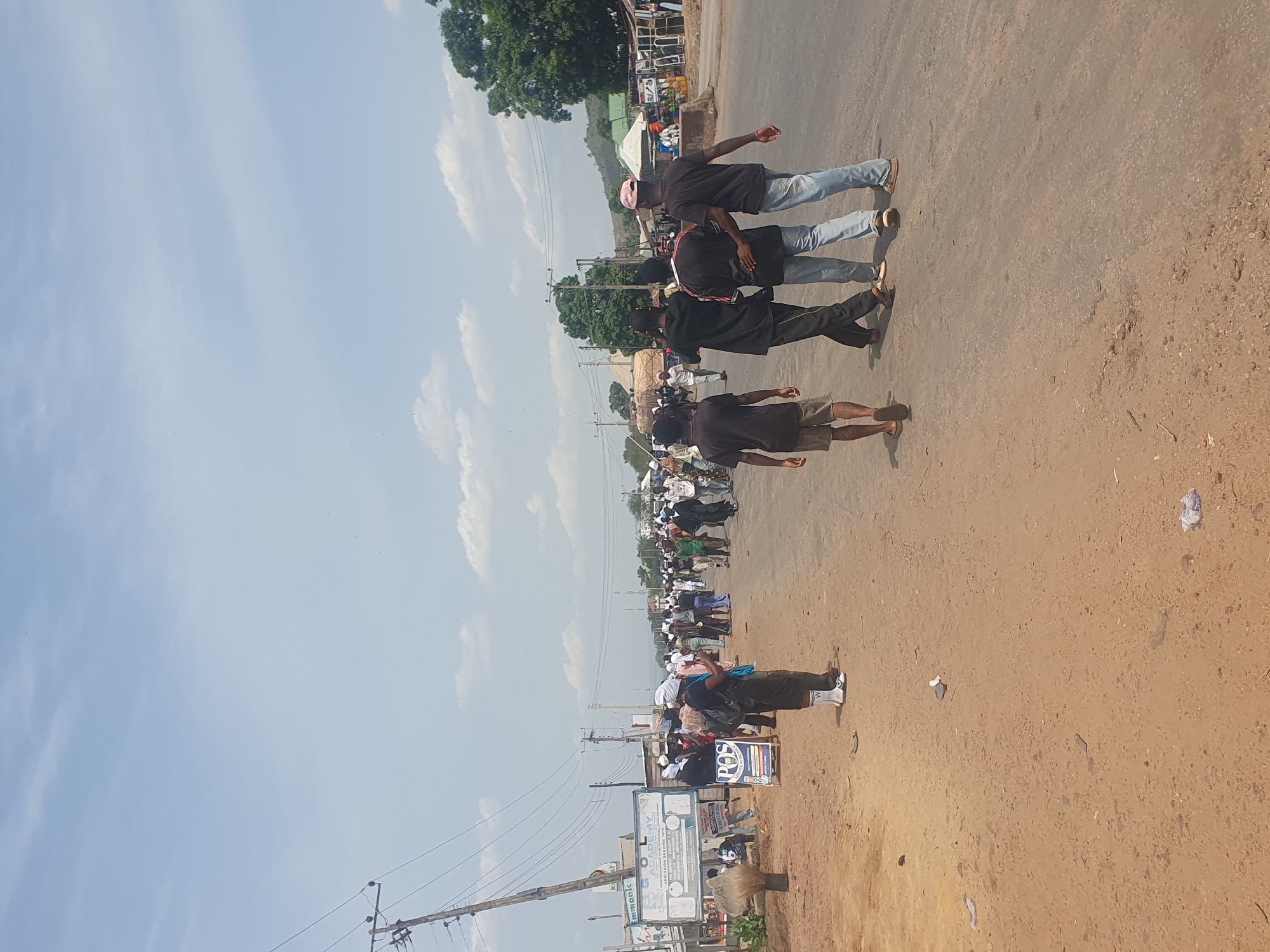 A street scene in Lokoja, Nigeria, shows a group of people walking or marching along a road or street. Several are wearing dark shirts and different types of pants. The road appears to be unpaved, with signage and utility poles visible along the side. There are other people present, and some are wearing white head coverings, suggesting possible religious attire. Other buildings and trees are visible in the background. The sky is blue with scattered clouds.