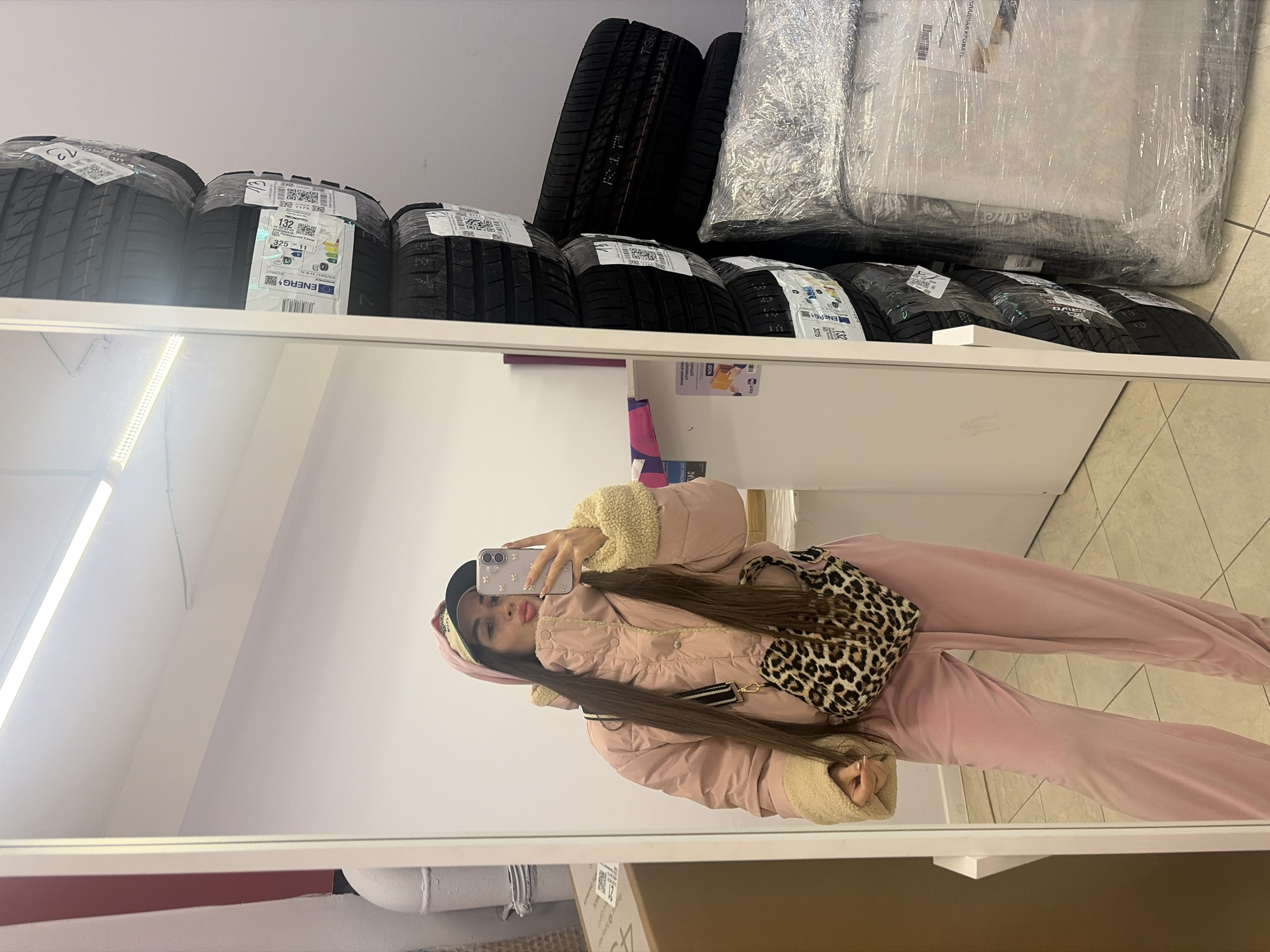 The scene is an indoor mirror selfie depicting a female subject in a commercial or storage environment in Saint Petersburg, Russia. The subject, a woman with long dark hair, is reflected in a large, white-framed mirror. She wears a light pink hooded puffer jacket with cream faux shearling cuffs, matching pink velour pants, and a black baseball cap under her hood. A leopard-print shoulder bag is slung across her body. She holds a light-colored smartphone with a floral case in her right hand, taking the photograph while pouting her lips.
To the right of the mirror, a vertical stack of at least six new car tires is visible, each wrapped in clear plastic and affixed with labels including European Tyre Labels and QR codes. Adjacent to the tires and on the floor, a large item identified by a partially visible Russian label as "СКЛАДНАЯ КРОВАТЬ" (folding bed) is wrapped in clear plastic film. The environment features white walls, light-colored tiled flooring, and an overhead LED strip light. A vertical white pipe and a brown cardboard box are partially visible in the far-left background. The Russian text on the packaging label is consistent with the location being in Russia, as specified by Saint Petersburg.