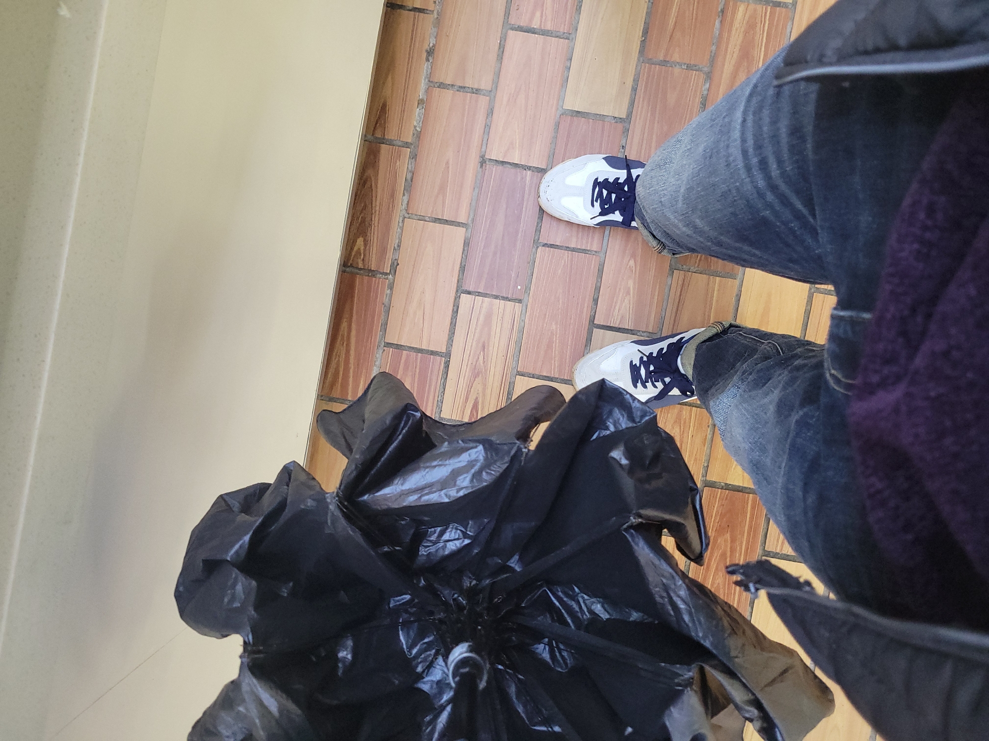 The image shows a person standing indoors on a brick-patterned tile floor. The person is wearing blue jeans and white sneakers with navy blue accents. A black umbrella lies collapsed on the floor next to the person's feet. A light-colored wall is visible on the left side of the frame. The person is wearing a black jacket and a purple sweater. The location is Unknown, Unknown.