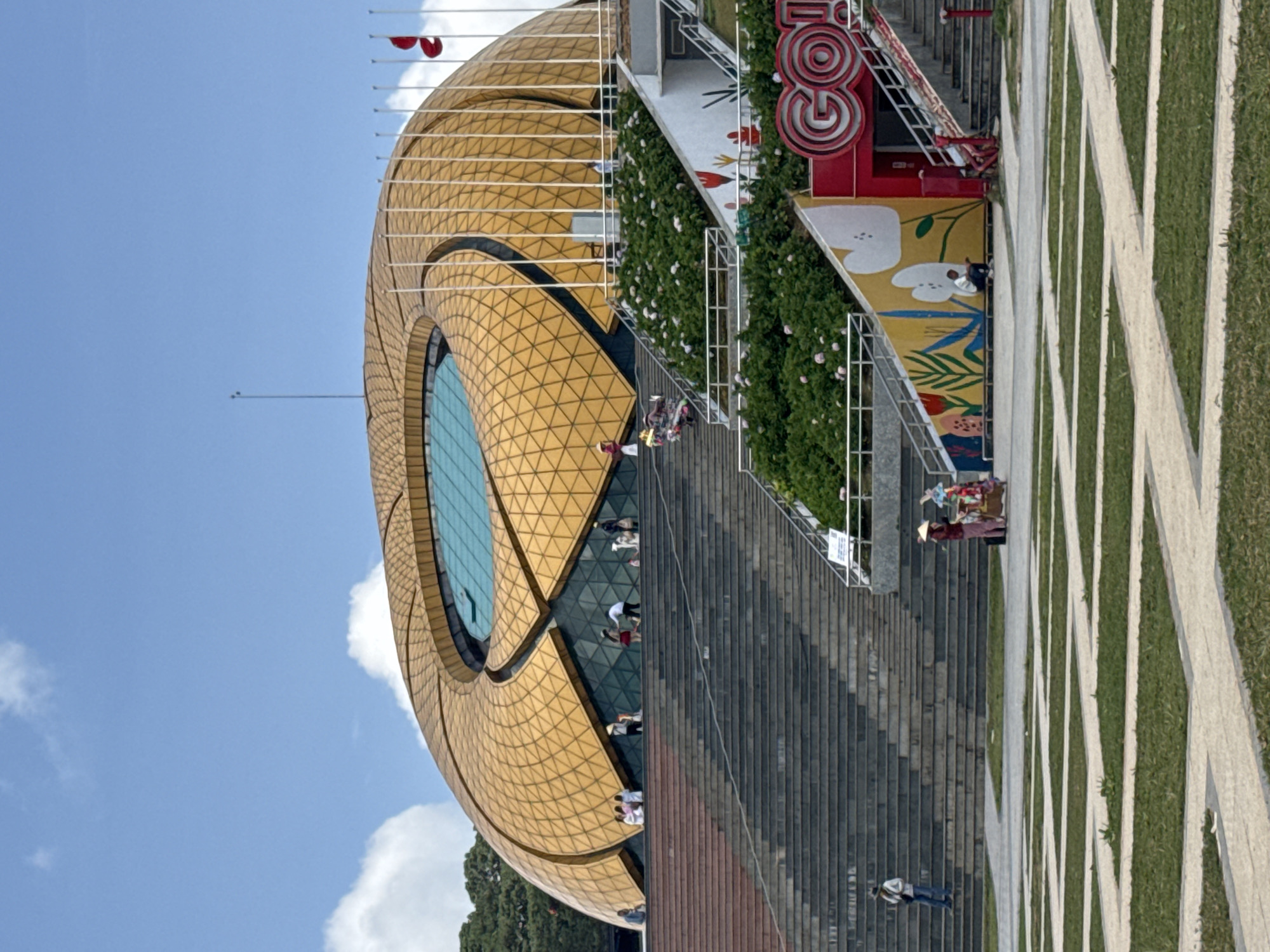 The image shows a wide shot of the Da Lat Cultural Palace in Da Lat, Vietnam. The building is characterized by its unique, golden, lotus-shaped roof. A grand staircase leads up to the entrance of the palace, with several people visible ascending and descending. The area surrounding the palace features lush green landscaping with flower beds. In the foreground, a patterned lawn with white borders is visible. To the right of the staircase, a section of wall is adorned with colorful floral murals. Several flags are planted in front of the building, and the sky is blue with scattered white clouds.
