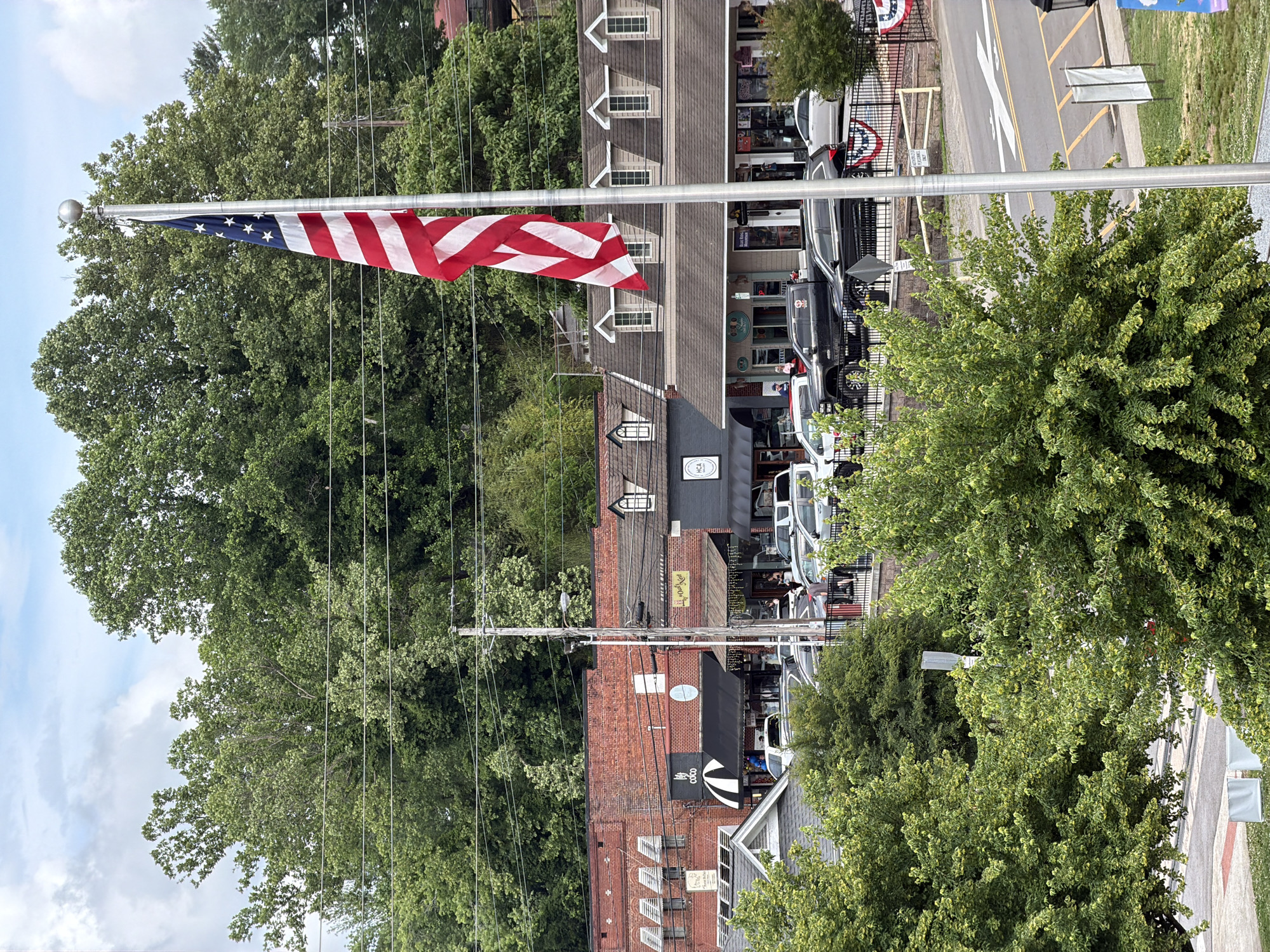 The image captures a street scene in Blue Ridge, Georgia, USA. An American flag flies prominently from a pole on the left side of the frame, with trees in the background. The street is lined with buildings, some brick and others wood-sided, featuring shops and businesses. Cars are parked along the street, and power lines are visible. A crosswalk marking is also shown.