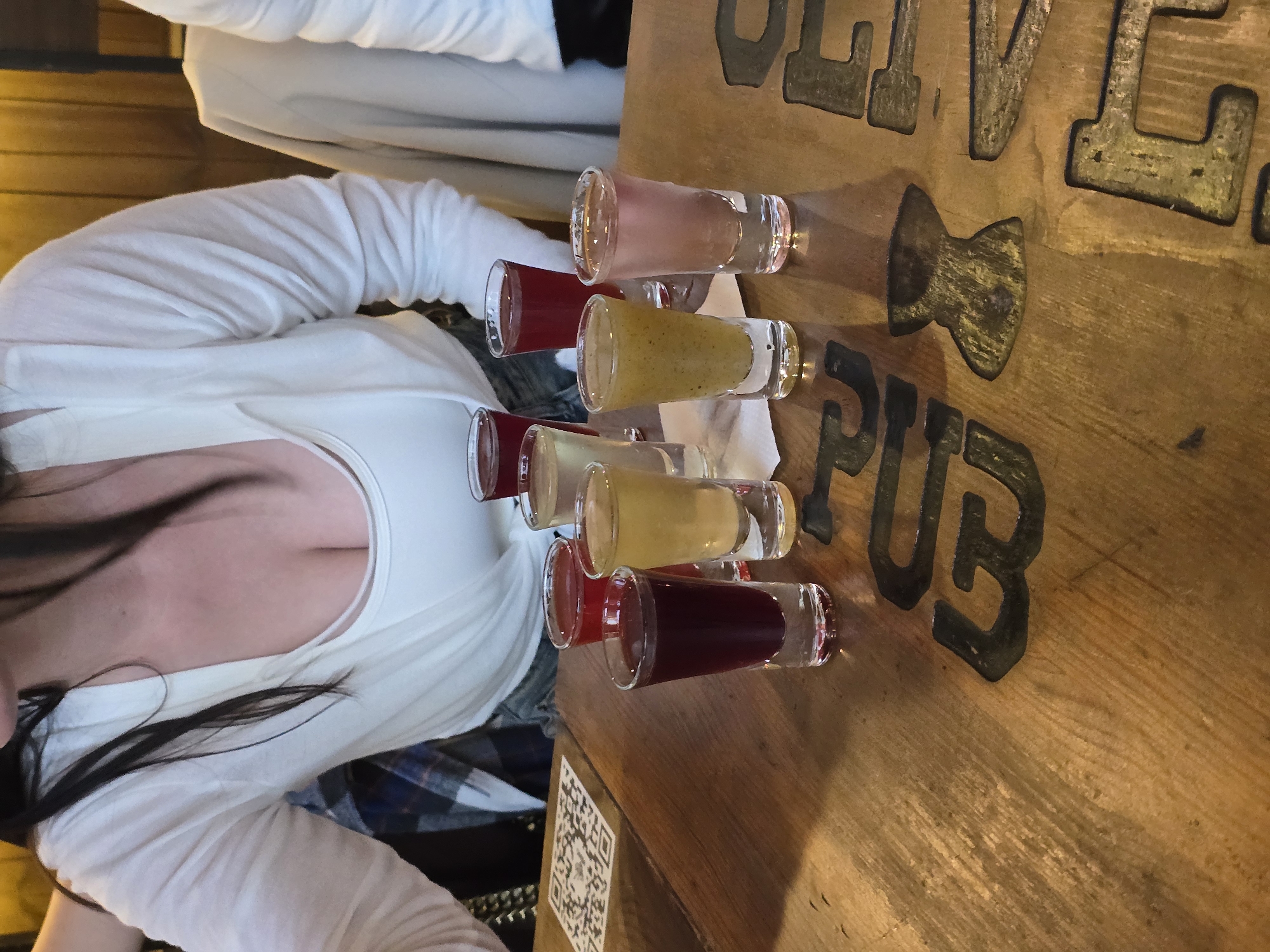 This image depicts an indoor scene at a pub or bar, identified as being in Vologda, Russia. The primary focus is on a wooden table laden with various colored shot glasses and the upper torso of a woman.
In the foreground, a sturdy, worn wooden table displays carved or inlaid dark letters, partially visible, spelling "PUB & ALE". Eight shot glasses containing different colored liquids are arranged on the table. From left to right, the visible shots include: one dark red/purple, one bright red, one yellow, one light yellow-green with visible particulates, one light pink, one yellow, one bright red, and one dark red/purple (partially obscured). Several of these glasses rest on small white rectangular paper napkins. A white square sticker with a black QR code is affixed to the table surface on the far left.
A woman is positioned behind the table, viewed from the mid-chest up. She has long dark hair, which falls over her shoulders. She wears a white long-sleeved, open-front top over a white tank top, revealing her cleavage. Her head is tilted slightly down, and her gaze appears directed towards the shots on the table. To her left, a plaid shirt (blue, white, and brown pattern) is visible, draped over a chair or worn by another person. To the right, the shoulder and arm of a second person, also wearing a white long-sleeved garment, are partially visible. A light beige or tan jacket hangs on a chair in the background to the right. The background features a warm-toned wooden wall or paneling, suggesting a rustic interior. The lighting is warm and ambient, characteristic of a pub setting.