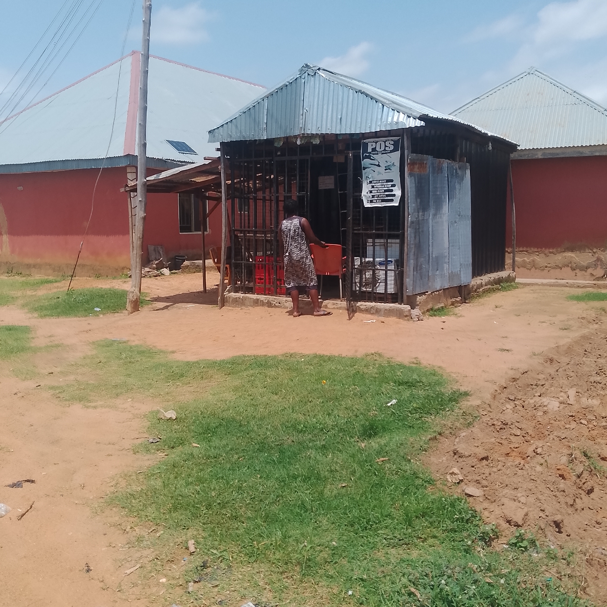 A person stands in front of a small, open-air kiosk with a corrugated metal roof. The kiosk appears to be a Point of Sale (POS) service provider, as indicated by a sign displaying "POS" prominently. The sign also lists services such as "Deposit Money," "Withdraw Money," "Send Money," "Pay Airtime," and "Pay Bills." Inside the kiosk, red crates of beverages are visible.
The scene is outdoors, with a dirt and grassy ground. The kiosk is situated next to a red building with a light-colored, possibly tin, roof. A utility pole with wires stands to the left of the kiosk. The sky is visible and appears to be partly cloudy, suggesting it is daytime. The overall setting seems to be a simple, practical commercial area. The person in the image is facing away from the camera, with their back towards the kiosk. They are wearing a patterned dress and flip-flops. The overall mood of the image is neutral, capturing a moment of everyday commercial activity.