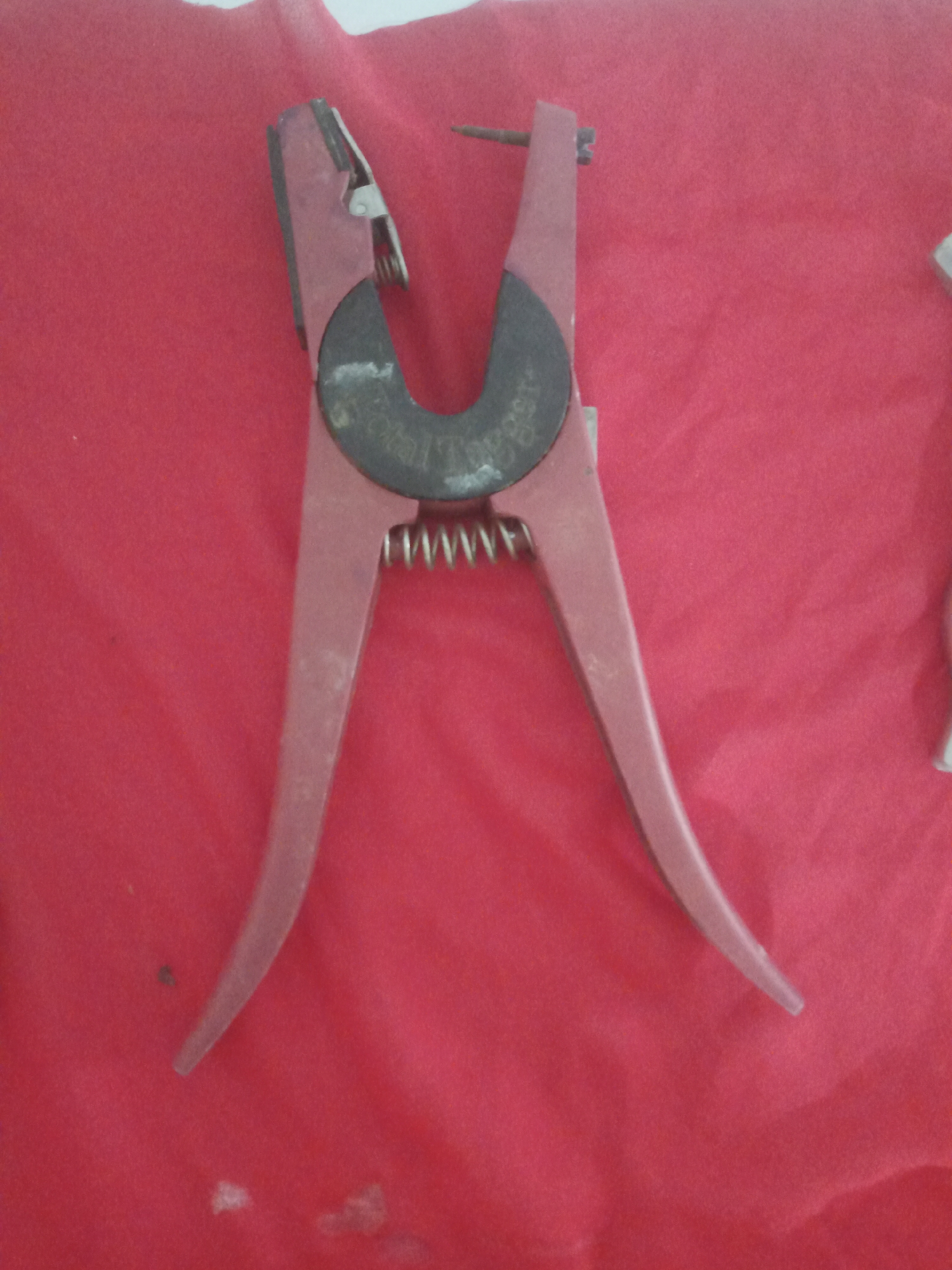 A pair of red ear tag pliers lies open on a red fabric surface. The pliers have metal components at the top, including a spring and a dark, circular piece with writing on it. The red handles of the pliers are wide and curve outwards. The fabric surface appears to be slightly wrinkled. The image does not contain any information about the city of La Tagua, Venezuela, or any people.