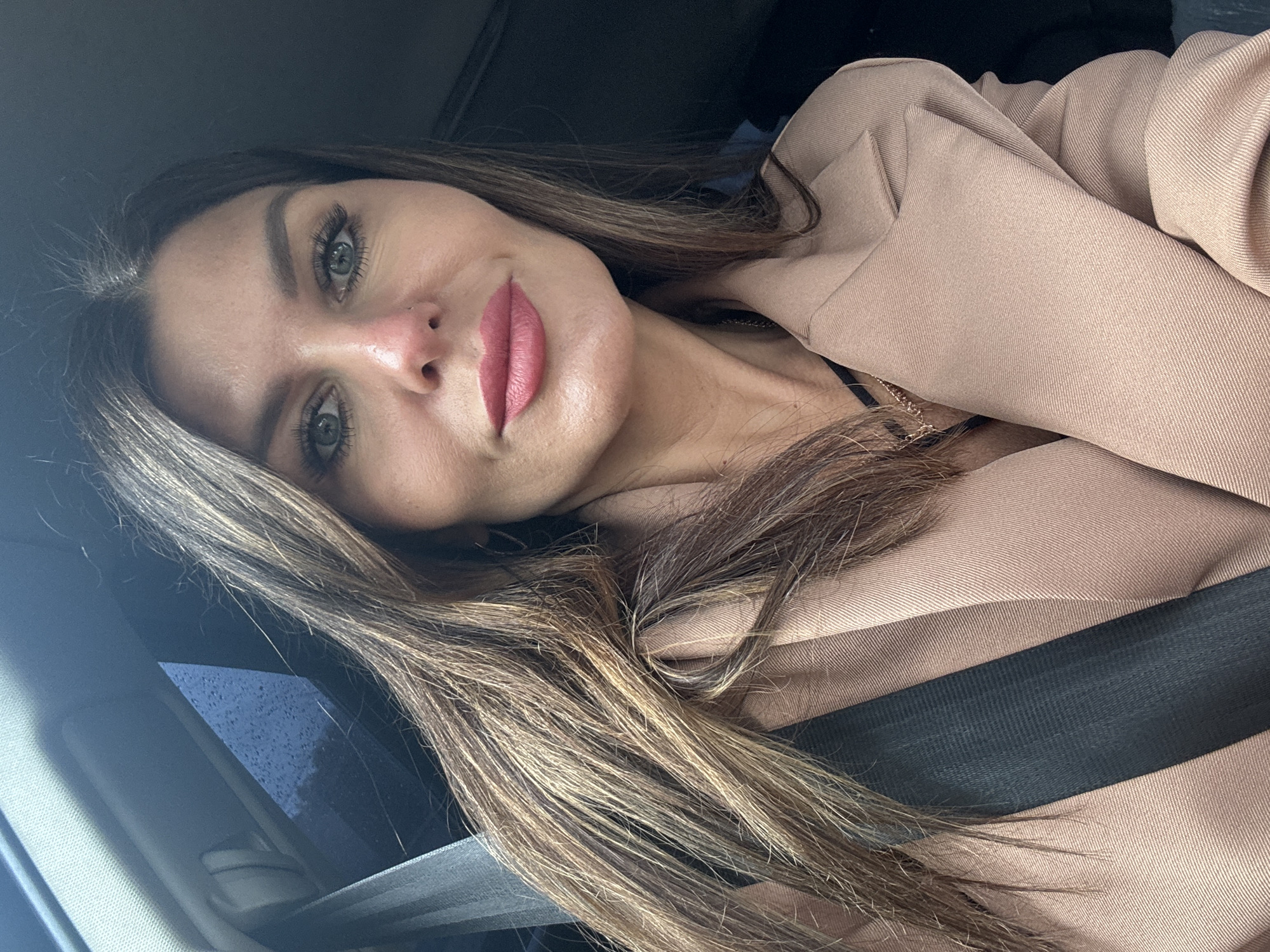 A female individual is captured in a self-portrait taken inside a car in Chaikovsky, Russia. She has long, wavy brown hair with lighter highlights, styled to fall over her shoulders. Her gray-green eyes are prominent, framed by long eyelashes and neatly groomed eyebrows. Her lips are full and adorned with a matte pink lipstick, curving into a gentle smile as she looks directly at the camera. A small nose piercing is visible.
She is dressed in a light beige or camel-colored blazer or jacket, worn over a dark garment, with a delicate gold-colored chain necklace visible around her neck. A black seatbelt is fastened across her chest and shoulder, indicating she is seated in the vehicle. The car's interior appears dark, with hints of a dark headliner and window. Through the window, a dark blue, textured external environment is visible, possibly suggesting an overcast sky or rain on the glass. The lighting is soft and natural, illuminating her face. No visible text is present in the image.