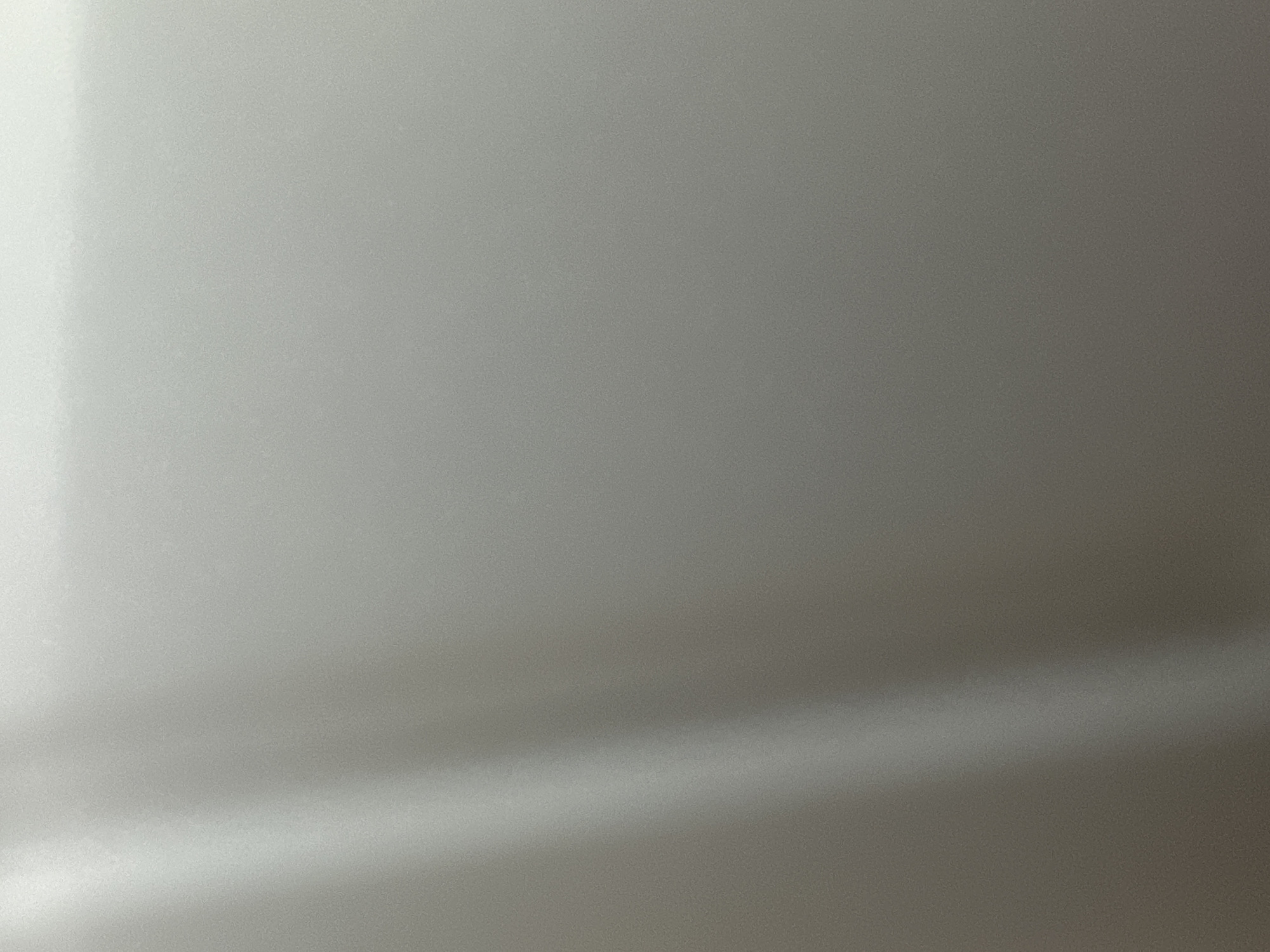 This is a close-up, abstract image of a textured, metallic surface, likely a wall or a vehicle. The surface is predominantly a cool, neutral gray with subtle variations in tone and highlights. There are no discernible subjects like people or objects. The lighting creates a soft gradient across the surface, with a bright, diffused illumination on the left side that fades into shadow on the right. A diagonal band of brighter light, possibly a reflection or a subtle curvature of the surface, cuts across the lower portion of the image. The texture appears to be a fine, granular pattern, giving the surface a slightly matte or brushed metal finish. There is no visible text, and the context of Belgrade, Serbia, does not provide any specific visual cues within the image itself. The time of day, weather, and any emotional context are not discernible from this abstract portrayal.