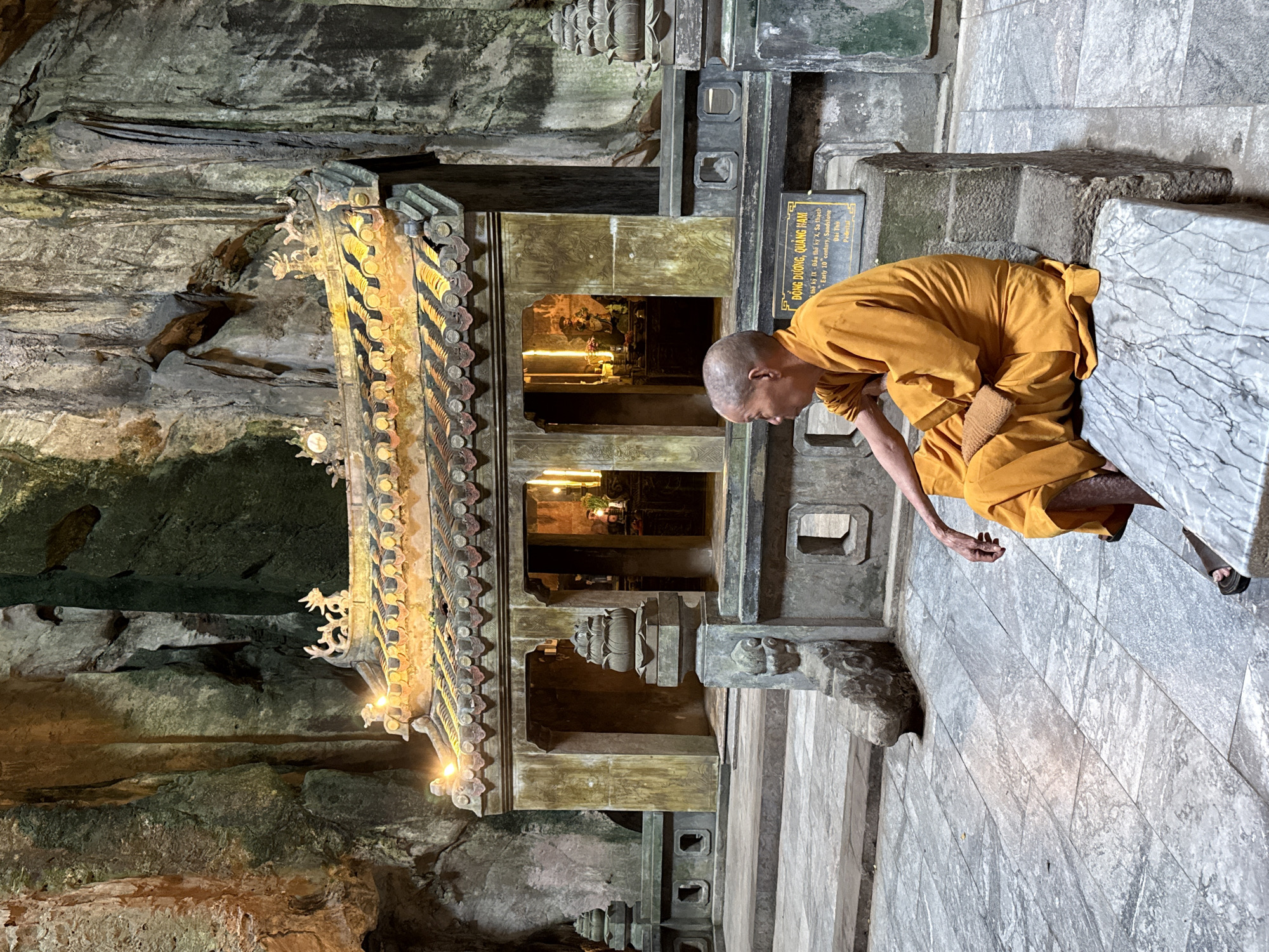 A Buddhist monk, identified as male, with a shaved head and wearing a saffron-orange robe, sits in profile on a marbled stone bench inside a large natural limestone cave. The location is within the Marble Mountains (Ngũ Hành Sơn) in Đà Nẵng, Vietnam. The monk faces left, with his right hand resting on his lap. Dark sandals are visible on his feet.
In the background, an ornate, weathered stone shrine or miniature temple structure is prominently featured, illuminated by warm artificial light. Its multi-tiered roof is covered with dark tiles and decorated with golden accents and stylized figures at the eaves and apex, from which small lights emit a yellow glow. The shrine has open archways, revealing multiple golden statues and glowing candles or lights within its interior. A low, stone balustrade with a decorative lotus-shaped finial on a pedestal partially encircles the shrine's raised platform.
To the right of the monk, adjacent to the shrine's base, a black rectangular sign with white text is visible, stating "ĐÔNG DƯƠNG, QUẢNG NAM / THẾ KỶ IX - ĐẦU THẾ KỶ X, Sa thạch / Early 10th century, Sandstone / Đại Thờ / Pedestal". The natural rock walls of the cave rise steeply in the background, displaying varied textures, striations, and colors ranging from grey to dark green, with patches illuminated by the shrine's light. The ground is paved with large, light-colored stone tiles.