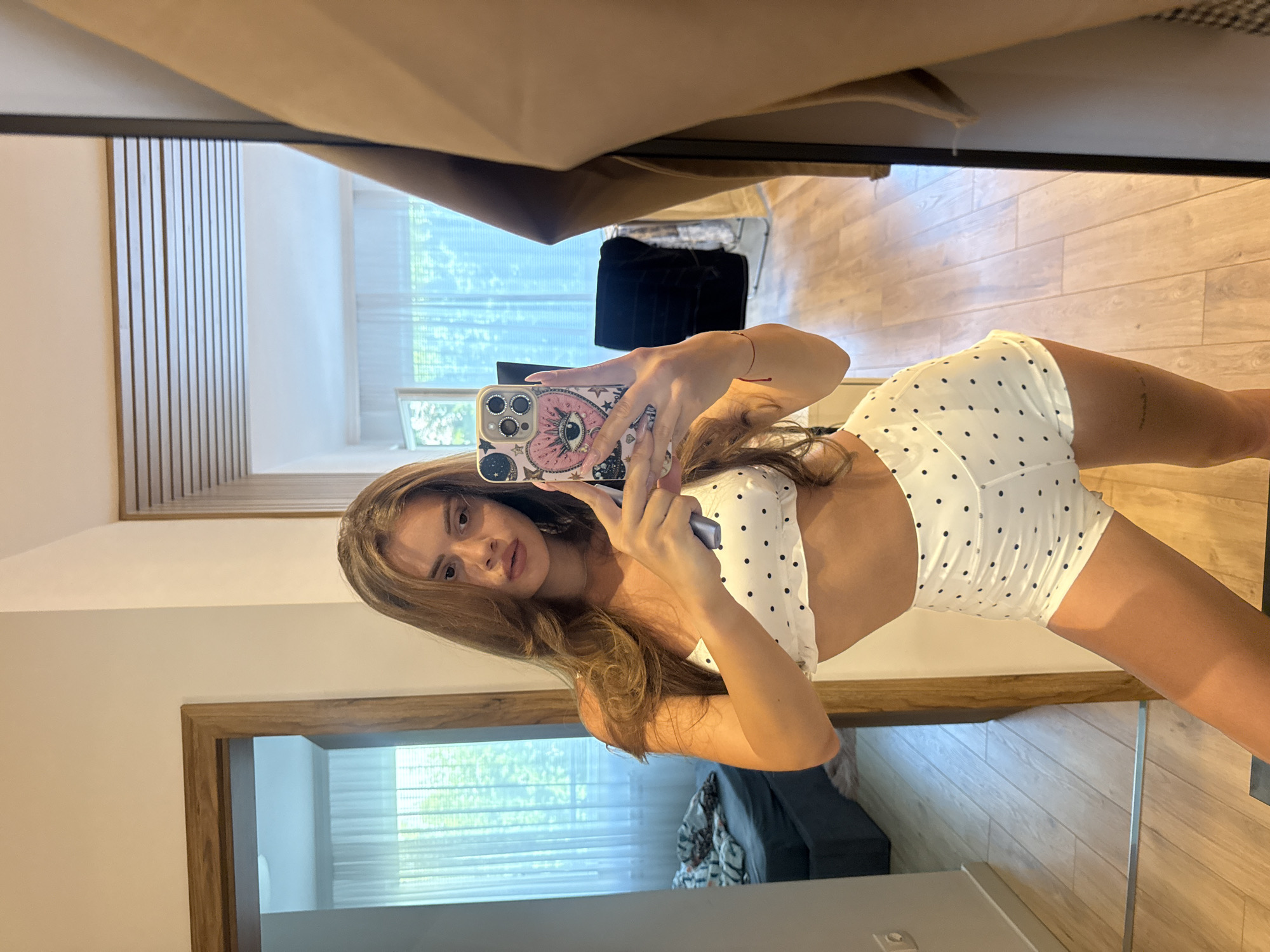A young woman with long, brown hair takes a selfie in a mirror. She is wearing a white polka dot bikini top and matching shorts. She holds a phone with a patterned case in her right hand. The background shows a bedroom with a bed and a window with sheer curtains. The room has light-colored wooden floors and a wooden door frame. This image was taken in a residential interior, likely a bedroom or dressing area, which could be located in the city of Plovdiv, Bulgaria, given its typical architectural and interior design elements often found in residential properties there.
