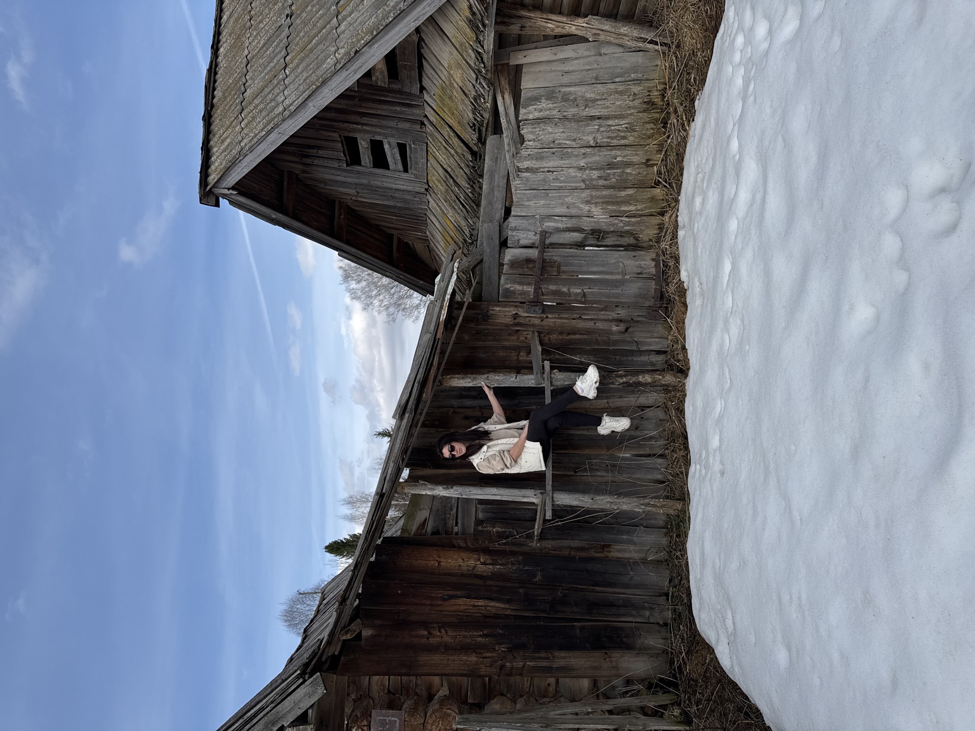 The photograph, taken in Вассята, Russia, depicts an individual suspended from a dilapidated wooden structure, possibly an abandoned building or shed. The person, wearing dark leggings, white athletic shoes, a light-colored top, and sunglasses, is situated within the decaying framework of the building. The structure itself exhibits significant weathering and disrepair, with missing or broken planks. A large pile of snow is visible adjacent to the building. The sky is partly cloudy.