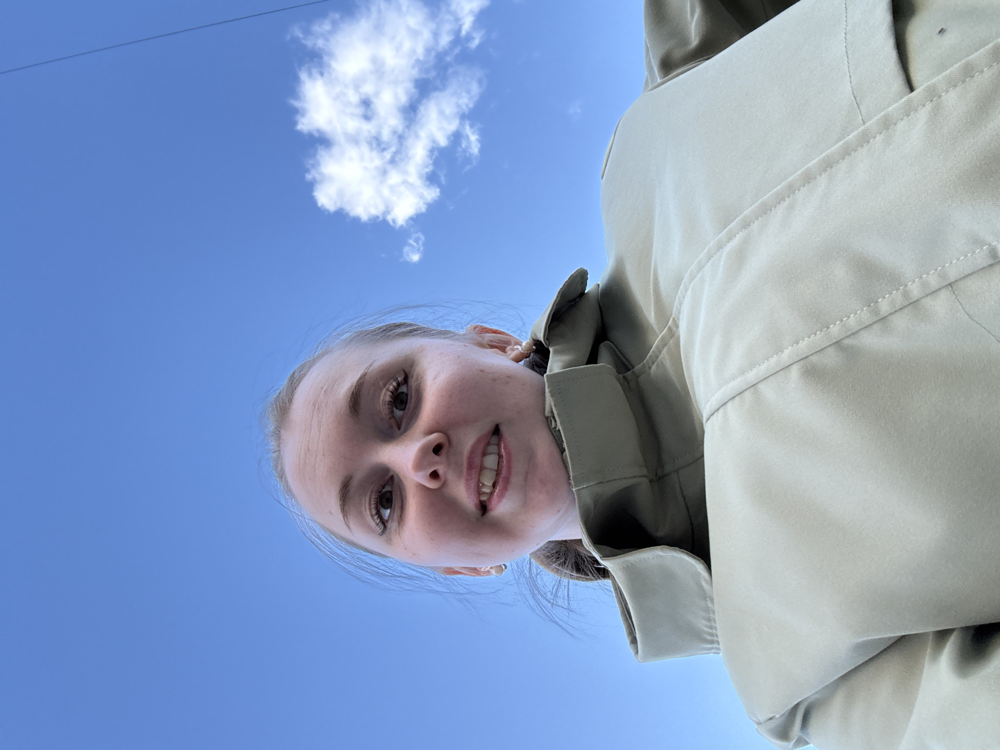 A young woman with light skin, brown eyes, and light brown hair smiles at the camera. She wears a light-colored jacket. Above her, a clear blue sky contains a small, puffy white cloud. The location is Unknown, Unknown.