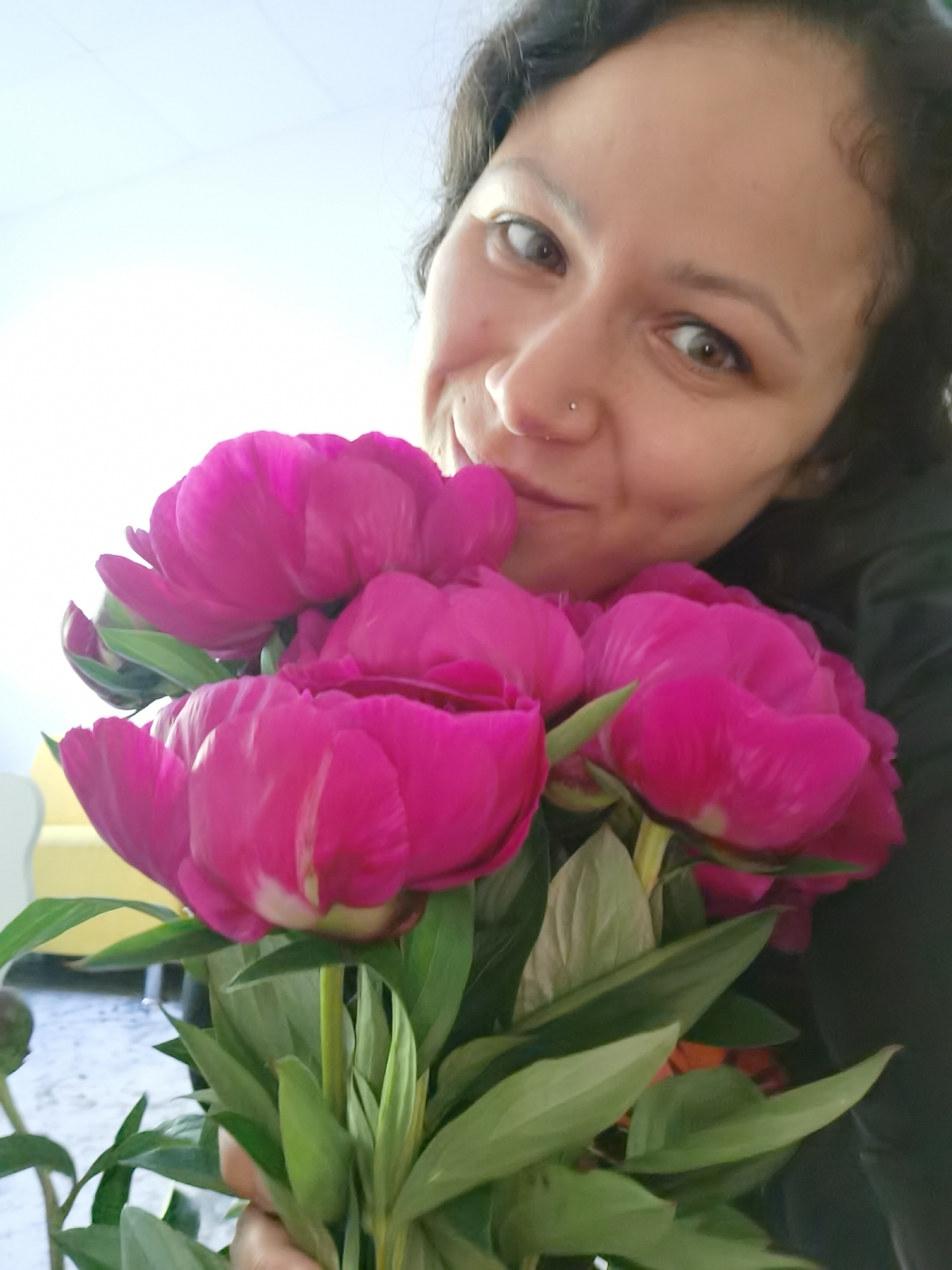 In this close-up, low-angle selfie, a woman with dark, curly hair and brown eyes is holding a bouquet of bright pink peonies. She is wearing a black hooded jacket. Her nose is adorned with a small silver stud. The background is a plain, bright white wall, suggesting an indoor setting. The focus is on the woman's face and the vibrant flowers, with the peonies occupying the majority of the frame. The lighting is bright, creating a soft glow. The specific location in the city of Chaikovsky, Russia is not determinable from the provided image.