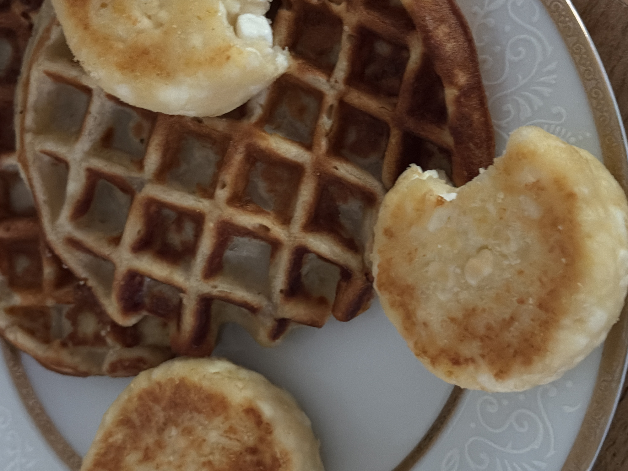 This is a close-up, overhead shot of a plate of food, featuring waffles and small, round, fried pastries. The plate has a decorative gold rim and a white, embossed pattern. The food is arranged on the plate, with a large waffle dominating the center and right side of the frame. Three of the fried pastries are visible. One is on the left side, another is on the top right, partially obscuring the waffle, and the third is in the bottom center, with a bite taken out of it. The pastries appear to be golden brown and slightly crisp on the outside, with a lighter, softer interior. The waffle is also golden brown and has a distinct grid pattern. The background is a wooden surface, slightly out of focus. The image does not contain any recognizable landmarks or elements that would indicate its location in Chirchik, Uzbekistan.