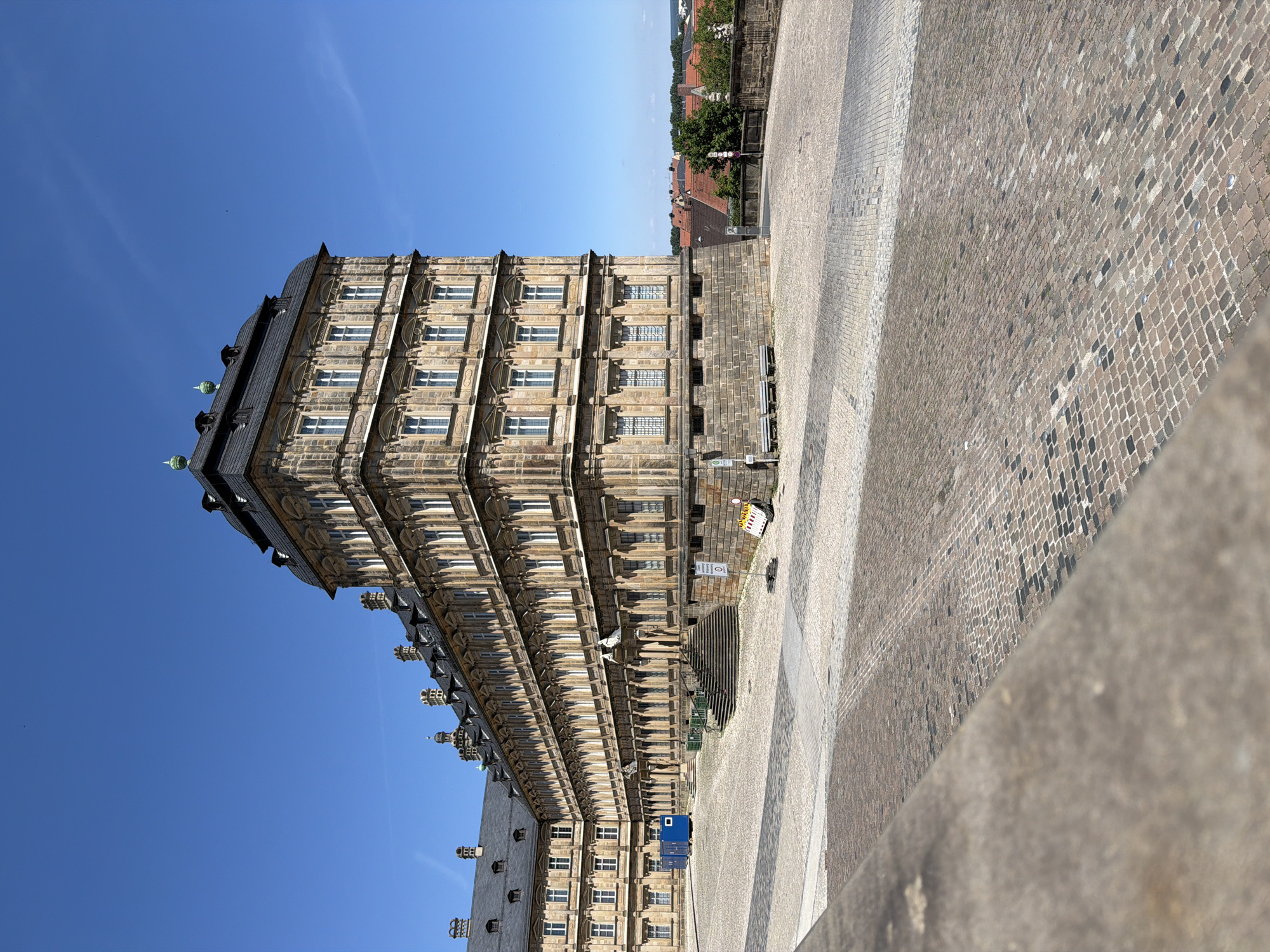 The image depicts the New Residence in Bamberg, Germany, under a clear blue sky. The grand Baroque palace, characterized by its extensive stone facade, multiple stories, and elaborate roofline with dormer windows and decorative elements, occupies a significant portion of the frame. In front of the palace, a large, paved courtyard, made of cobblestones in various patterns and colors, stretches out. To the left of the palace, a blue portable building is visible. Two park benches are situated against the stone base of the palace. Beyond the palace, the rooftops of the city of Bamberg, with their characteristic red tiles, are visible, along with some trees and greenery. The scene is tranquil, with no people visible, suggesting a peaceful daytime ambiance.