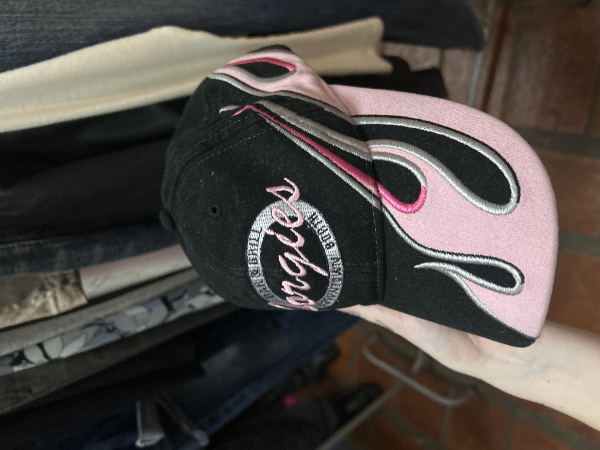 A black baseball cap with a light pink bill is being held in a person's hand. The cap has a logo embroidered on the side, featuring the name "George's" and the words "BAR & GRILL" within a circular design. The bill of the cap is light pink and has a flame-like design outlined in light grey and pink. The background shows a stack of clothing items, and a glimpse of a brick wall or floor. The city of Buenos Aires, Argentina is not discernible from this image.