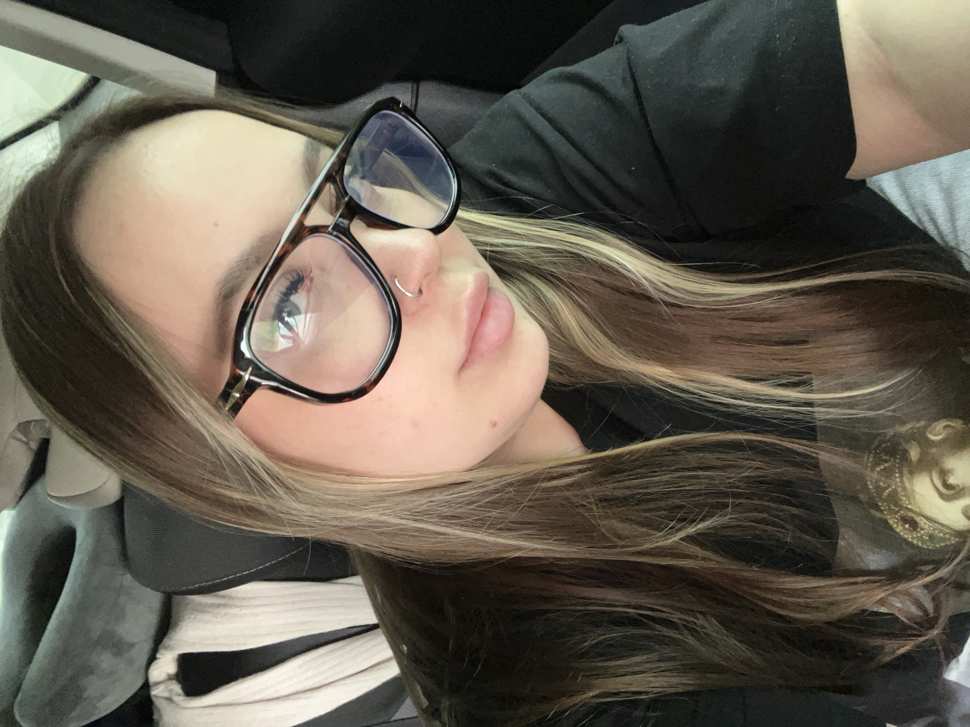 A close-up, slightly angled portrait captures a young individual with light-to-medium brown hair featuring blonde/caramel highlights. The subject's head is tilted to their left, with their face primarily on the left side of the frame. They wear large, dark tortoiseshell-framed glasses that cover part of their left eye, and a small silver hoop nose ring is visible in their left nostril. The person's lips are slightly parted, and their expression appears neutral and calm. They are wearing a black top.
The background is softly blurred, showing dark, upholstered surfaces, suggesting an indoor or enclosed setting, possibly a vehicle interior. In the lower left, beneath the person's hair, a light-colored, ribbed fabric is visible next to some grey material. On the right side, partially obscured by their long hair, a glimpse of a gold-colored, ornate object, possibly a decorative item or part of a doll, can be seen. The lighting is soft and ambient, offering no distinct cues for time of day or weather. There is no visible text in the image.