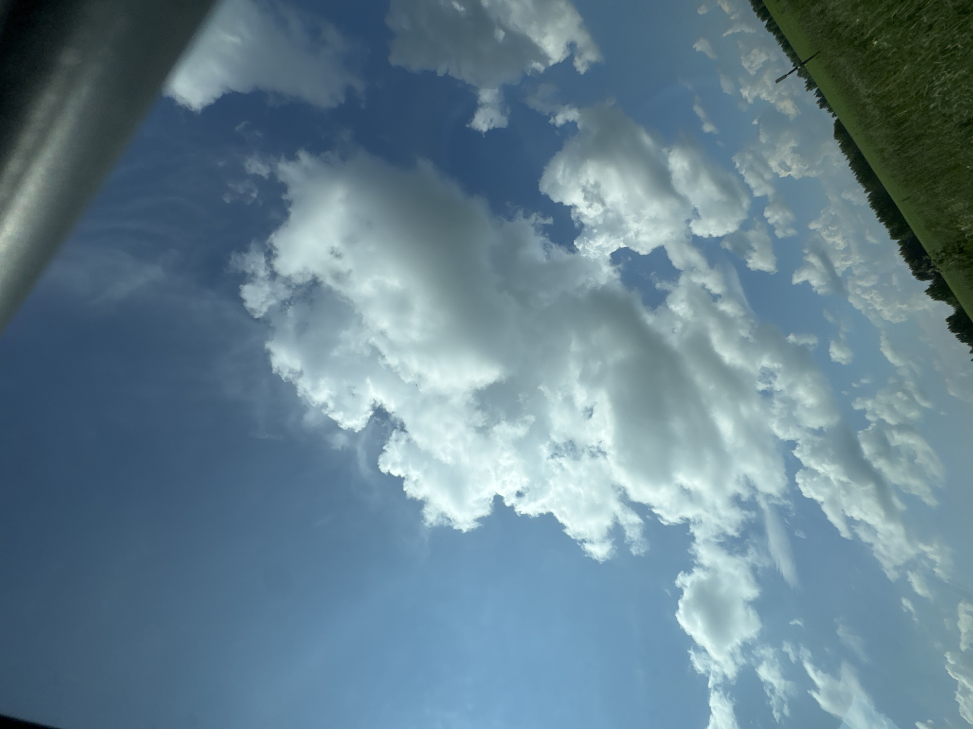 The image is a landscape photograph taken from a low angle, capturing a vast expanse of blue sky dotted with white, fluffy cumulus clouds. The sky takes up most of the frame, with a hint of green grassy fields and a line of trees visible along the bottom edge, suggesting a rural or semi-rural setting. A dark, metallic object, possibly part of a vehicle or structure, intrudes into the upper right corner of the image, casting a shadow or obscuring a portion of the sky. The overall impression is one of a clear, bright day, likely during daylight hours, given the illumination of the clouds. There are no people or discernible actions taking place. The image does not provide any information about the specific location within Vassyata, Russia, or any identifiable landmarks.