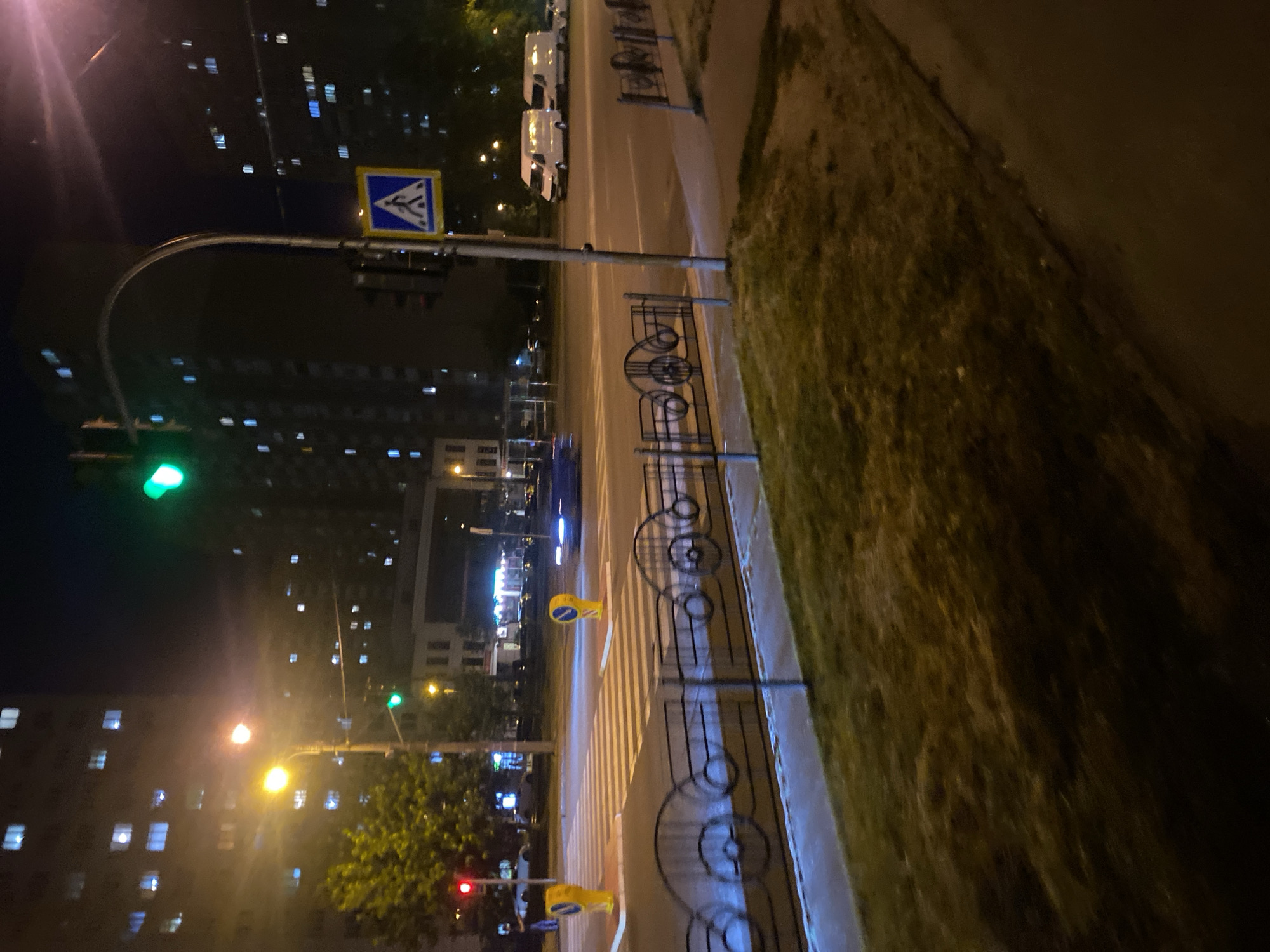 This image depicts an urban street scene at night in Kyiv, Ukraine. A pedestrian crossing is illuminated by traffic signals and streetlights. Two yellow bollards are positioned at the sides of the crossing. In the background, tall apartment buildings with illuminated windows dominate the skyline. A traffic light above the crossing shows a green signal. A pedestrian crossing sign with a blue background and a white pedestrian symbol is visible on a pole. Several white vans are parked on the right side of the road. A dark-colored vehicle is blurred in motion, suggesting it is driving across the intersection. A decorative metal fence with circular patterns runs along the sidewalk in the foreground, separating the street from a grassy area. The overall ambiance is that of a typical city night, with the artificial lights of buildings and vehicles creating a dynamic and somewhat hazy atmosphere.