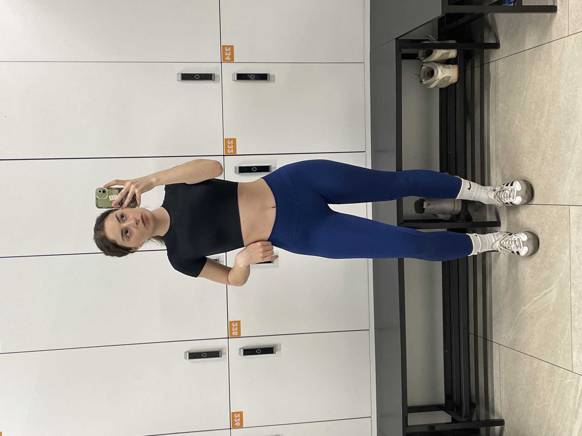 A young woman is standing indoors, likely in a gym locker room, taking a selfie in front of a row of white lockers. She is wearing a short-sleeved black crop top and blue leggings, with white socks and athletic shoes. She is holding a smartphone in her left hand. A metal bench and a shoe rack are positioned to the right of the lockers. There is no indication of the city of Moscow, Russia, in the image.