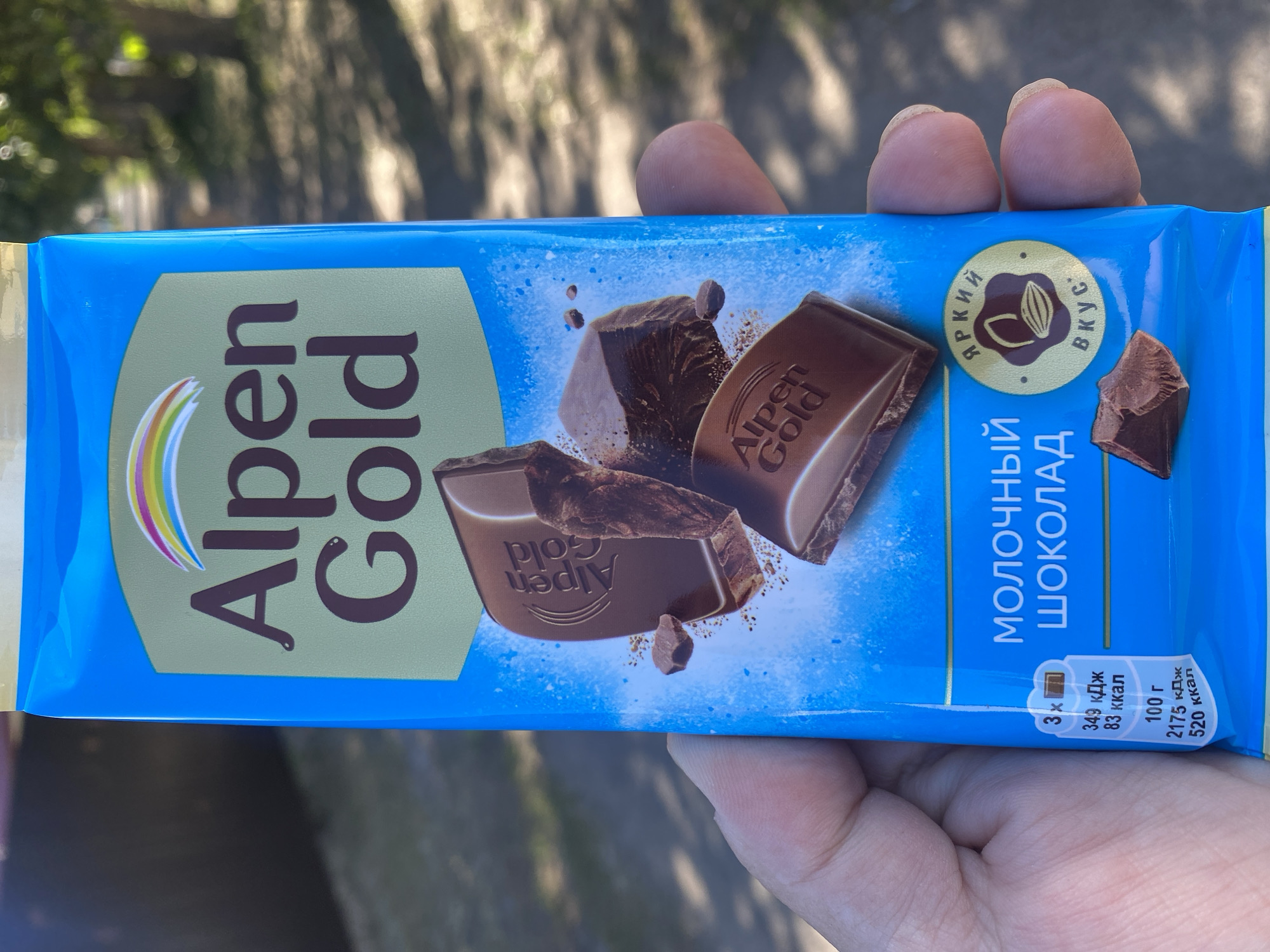 The image shows a person holding a bar of Alpen Gold milk chocolate. The wrapper is primarily bright blue with a light green oval containing the Alpen Gold logo and the text "Alpen Gold" in brown lettering. Below this, an image of several broken pieces of chocolate is depicted against a blue, abstract background. To the left of the chocolate pieces, there is a nutritional information section. To the right, a circular emblem with the text "Яркий вкус" (Bright taste) is visible. The background of the image is blurred, showing a sunlit path or sidewalk lined with trees, suggesting an outdoor setting. The person's hand, with visible fingerprints and fingernails, is holding the chocolate bar. The location is identified as Mamontovo, Russia, but there are no specific visual cues in the image that pinpoint this exact location within the city.