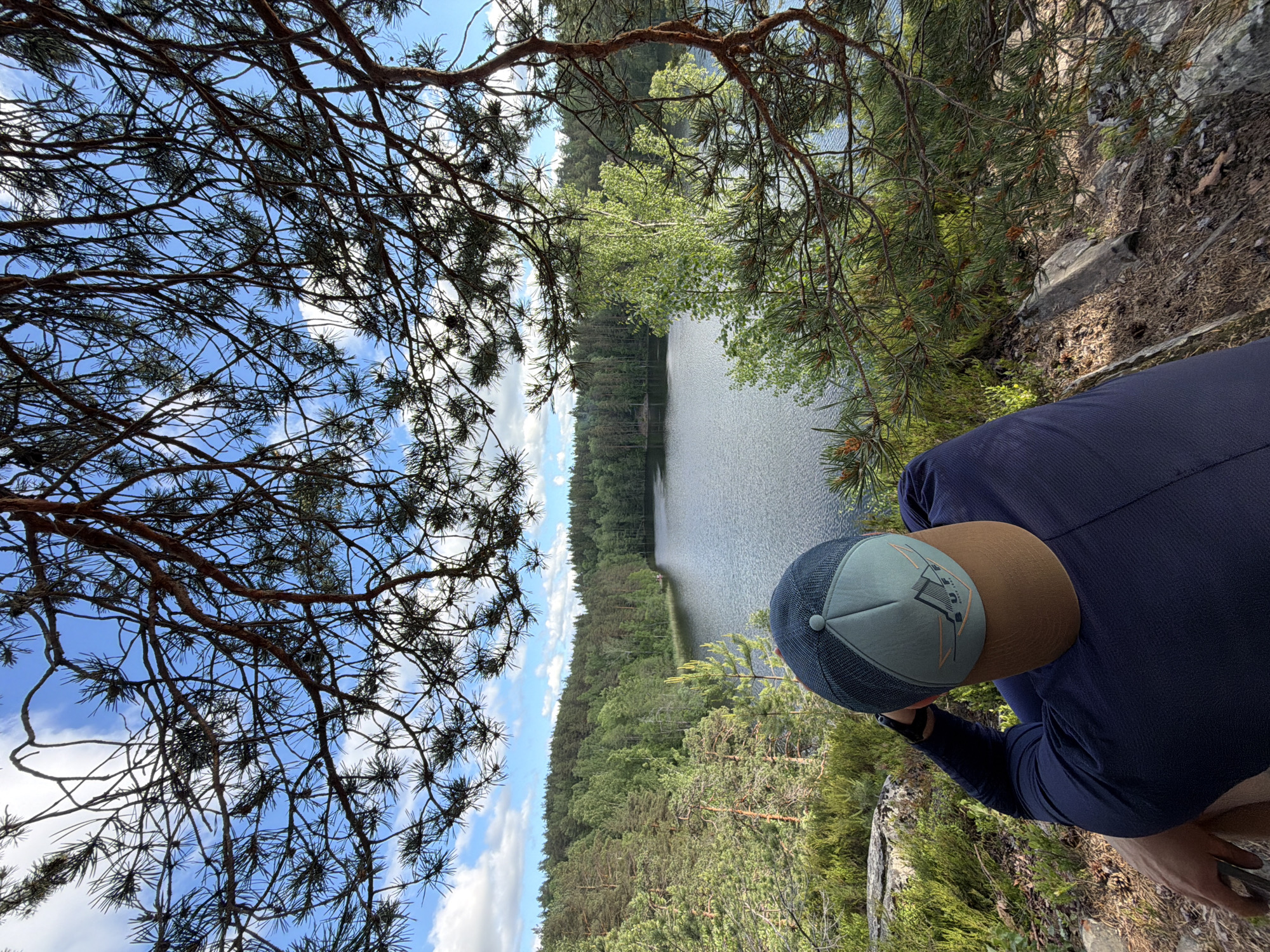 A person wearing a navy blue long-sleeved shirt and a baseball cap is seated on a rocky outcrop, looking out at a lake. The cap is teal and tan with an emblem. The lake is surrounded by a dense forest of pine and deciduous trees under a bright, cloudy sky. The image is taken from a high vantage point, looking down over the person and the landscape. The location is not identified as being in the city of Ruch'i, Russia.