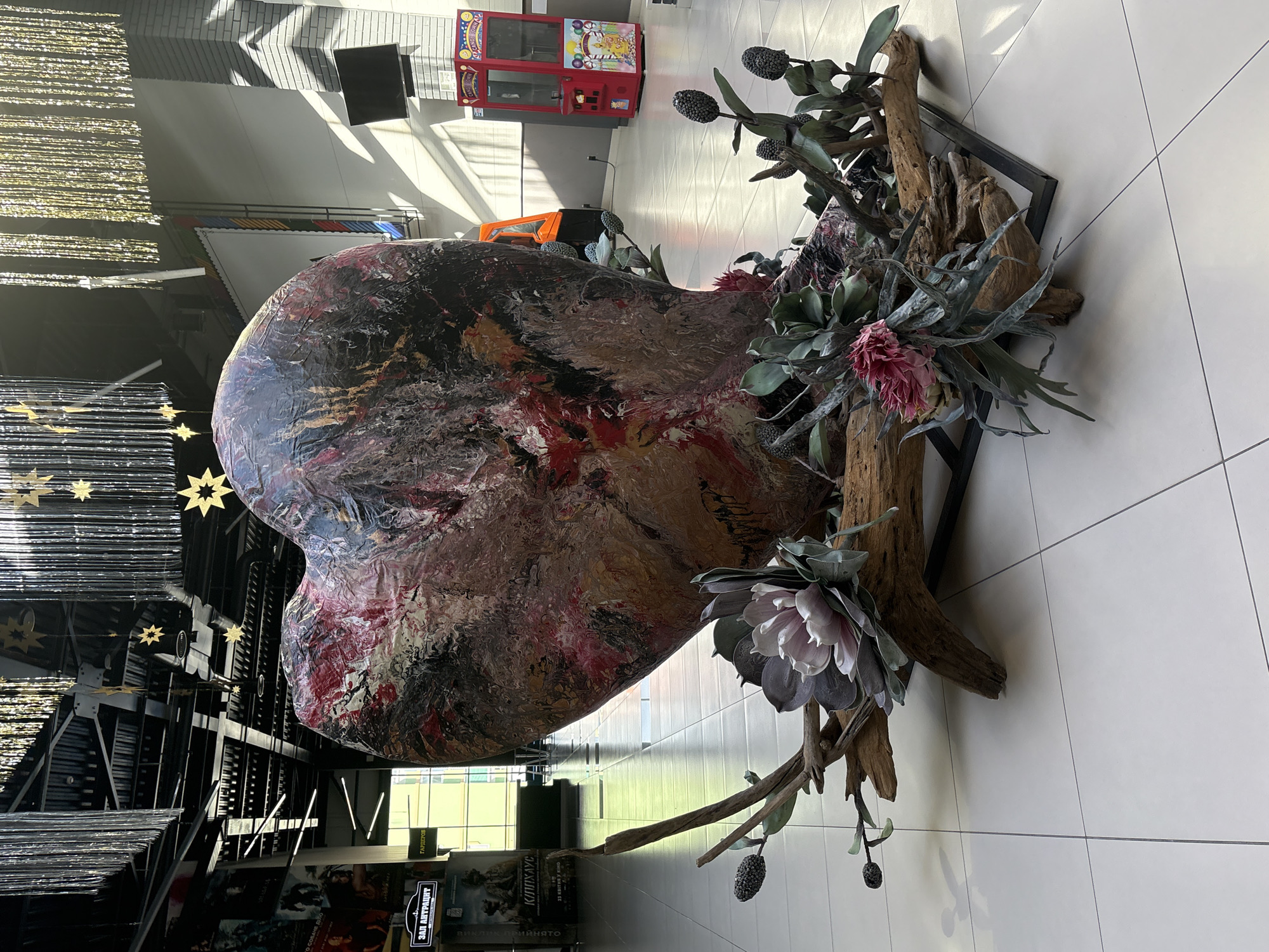 The image depicts an indoor space in what appears to be a commercial or entertainment venue in Hatne, Ukraine. The focal point is a large, abstract heart sculpture resting on a wooden structure and decorated with artificial flowers and foliage. The sculpture is painted with a mixture of colors, including brown, red, and black. To the left of the sculpture is a dark, decorative installation with hanging metallic strands and star-shaped accents. Further back, there are movie posters and signage in what appears to be a cinema or theater area. In the background, there is a bright, tiled floor, and a red vending machine is visible near a sunlit window.