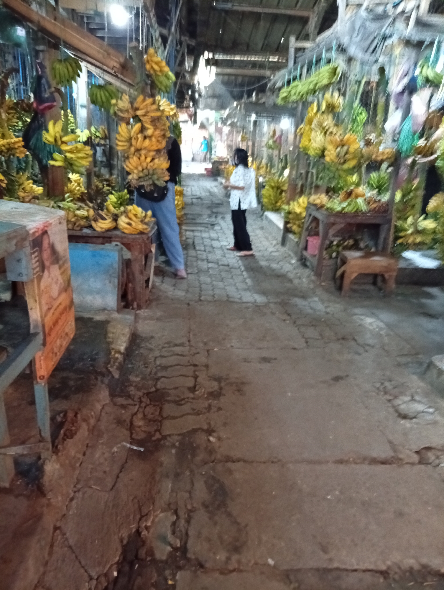 A bustling outdoor market in Larangan, Indonesia, is depicted with numerous stalls laden with ripe bananas. The ground is a rough, paved surface showing signs of wear and tear, with cracks and unevenness. Several people are present, including a woman in a white shirt and dark pants standing near a stall of yellow bananas, and another person in the background wearing a blue shirt. Various bunches of bananas, both green and yellow, are prominently displayed, hanging from wooden structures and resting on tables. The market appears to be covered by a corrugated metal roof in some areas, suggesting an open-air structure with some overhead protection. The scene is busy and vibrant, indicative of a typical market day.