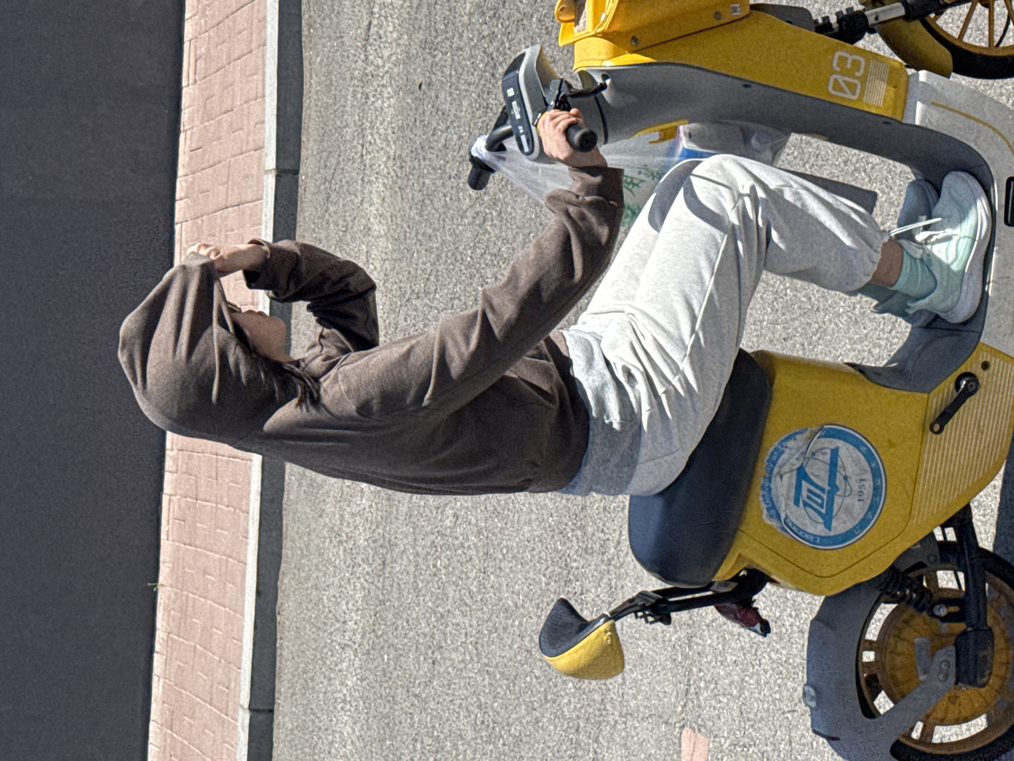 The image, rotated 90 degrees counter-clockwise, shows a person sitting on a yellow and gray shared electric scooter or e-bike. The scene is outdoors on what appears to be an asphalt road or pavement, next to a vertical brick-patterned wall.
The subject is wearing a brown hooded sweatshirt with the hood pulled up, light gray sweatpants, light blue sneakers, and light blue socks. They are holding the scooter's handlebars with their right hand. Their left hand is raised, shielding their eyes from what appears to be bright sunlight, indicating a sunny day.
The shared scooter is mainly yellow with gray components, including a gray section at the front displaying the number "03." A circular blue and white sticker is affixed to the scooter's side, featuring text that partially reads "UNIVERSITY OF LIBERIA" at the top and "1951" at the bottom right. The scooter has pedals and a small black seat. The bright, clear lighting suggests it is daytime. The setting appears to be an urban or suburban environment, consistent with Zhongtun, China.