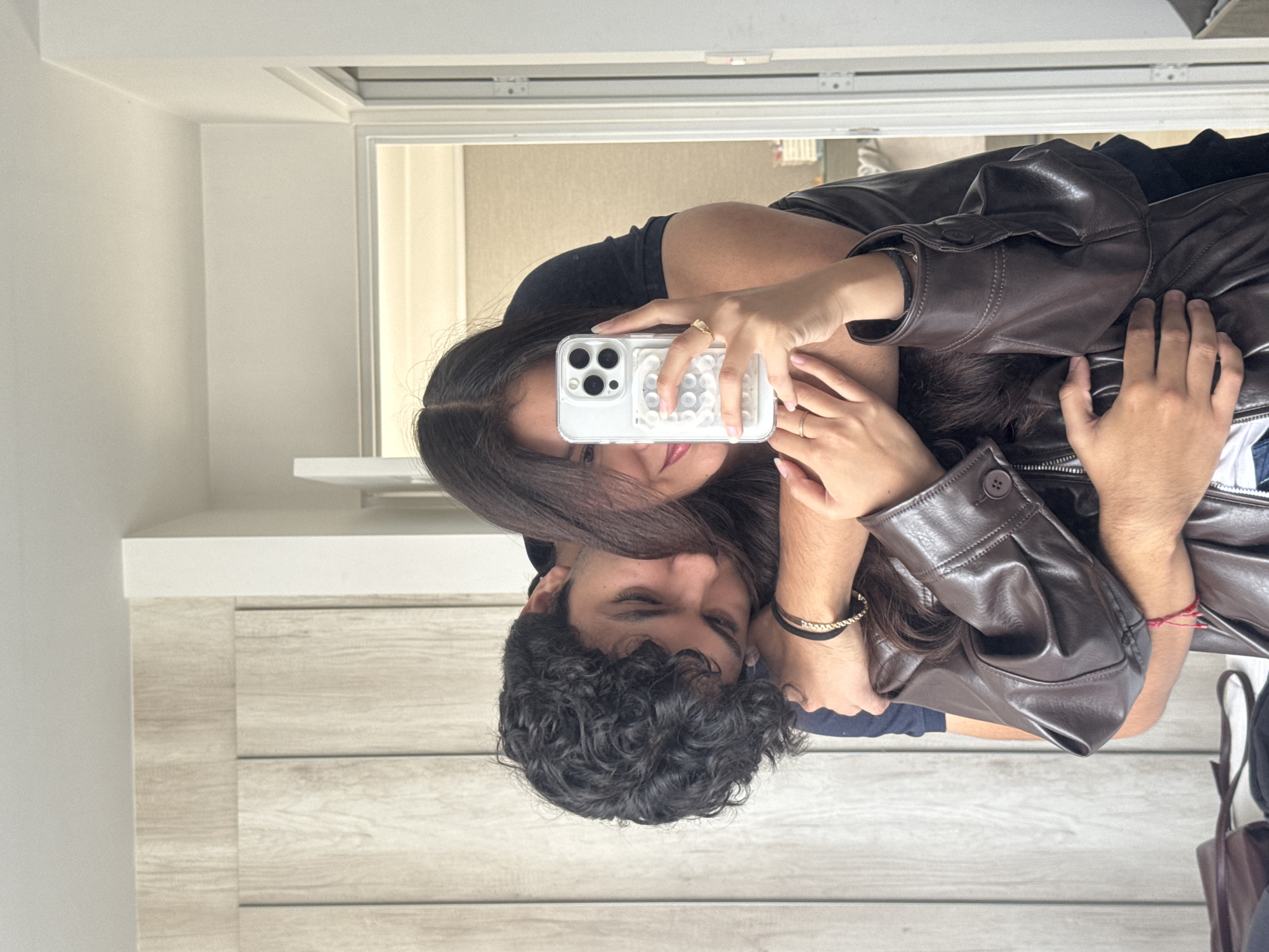A selfie of a couple in a room with light gray horizontal plank wall paneling, reflected in a mirror. The woman has long dark hair and is wearing a black top. She is holding a white iPhone with a clear case. The man has curly dark hair. The woman is wearing a brown leather jacket. The location cannot be determined, nor can a connection to San Francisco Ocotlán (Ocotlán), Mexico.