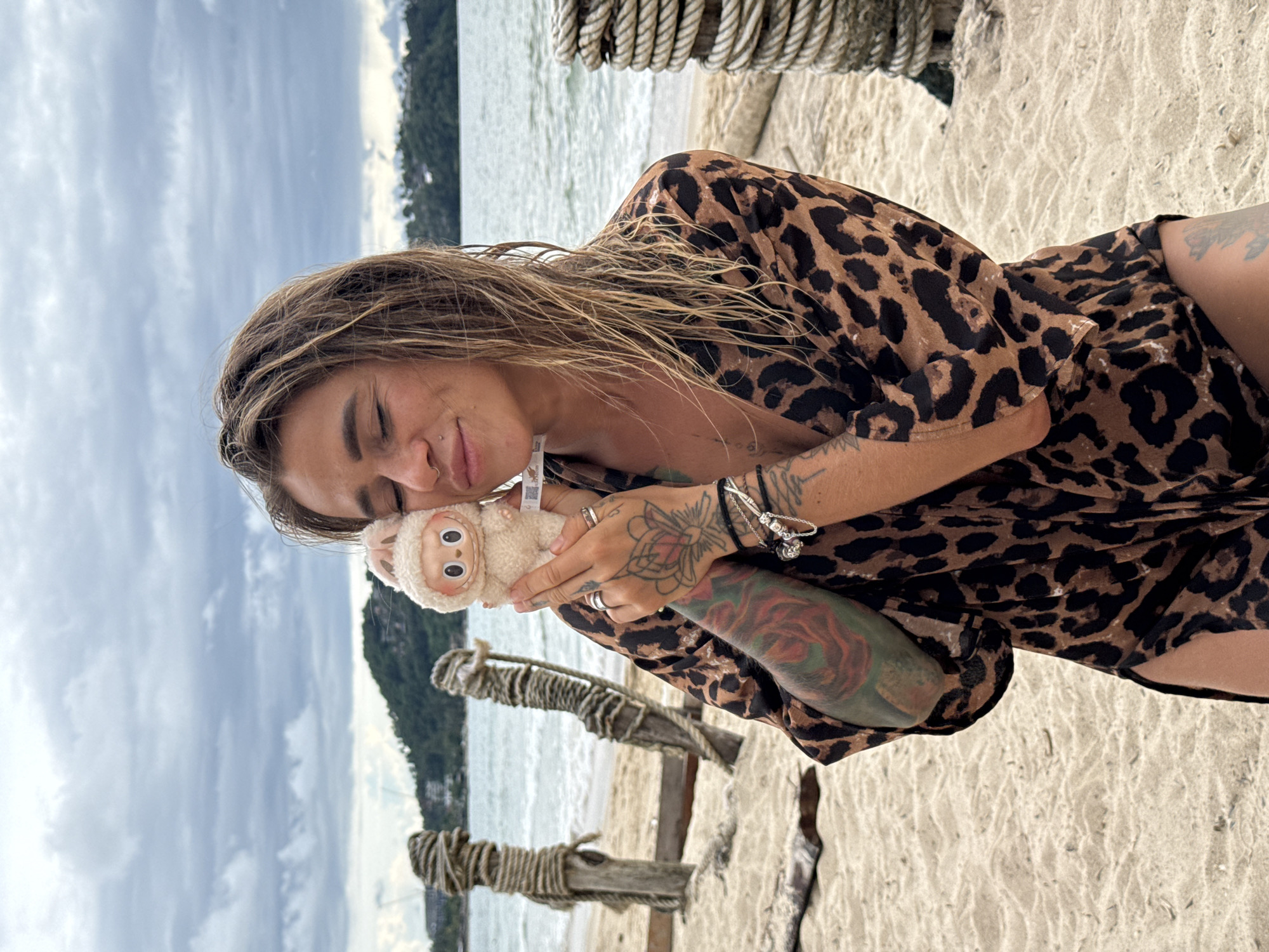 A woman with wet, blonde hair and closed eyes holds a small, plush toy with pink ears and a white body. She is wearing a leopard-print dress and several bracelets and rings. The woman has tattoos on her arms and leg. She is standing on a sandy beach with the ocean and a green, tree-covered hill in the background. There are wooden posts wrapped in rope on the left side of the image and a stacked rope post on the right. The sky is overcast. The location is Choeng Thale, Thailand.
