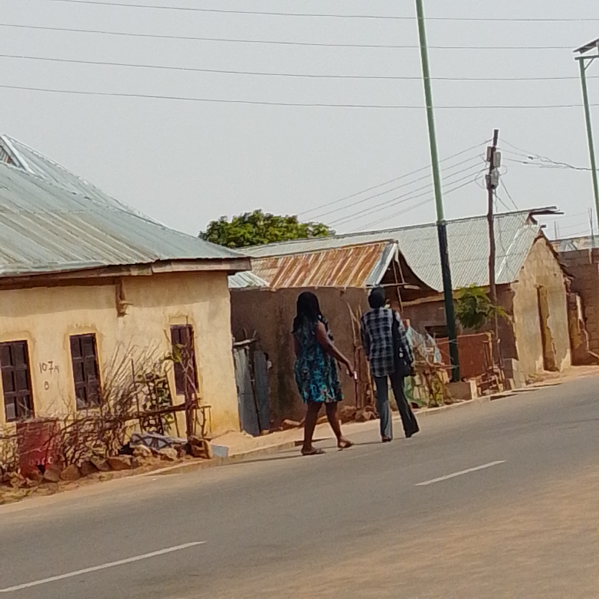 The image depicts a street scene in Gombe, Nigeria, likely in a residential or mixed-use area. In the foreground, a paved road with a white lane marking runs diagonally from the bottom left to the bottom right. On the left side of the road, a single-story building with a corrugated metal roof and beige stucco walls is visible. The building has dark-framed windows and the number "107A" is spray-painted on its facade. The area in front of this building is somewhat unkempt, with rocks and dry vegetation.
Further down the street, on the right side of the road, are several other structures with corrugated metal roofs, some appearing older and more weathered than the first building. One of these structures has a prominent entrance.
Two individuals are walking on the sidewalk adjacent to the road, moving away from the camera. The person on the left is wearing a dark, floral-patterned dress and sandals. The person on the right is wearing a checkered shirt and dark trousers. They are walking in the same direction.
In the background, overhead power lines are visible against a hazy sky. Green foliage from trees can be seen behind the buildings. The overall impression is that of a moderately developed urban or semi-urban environment in Gombe, Nigeria.