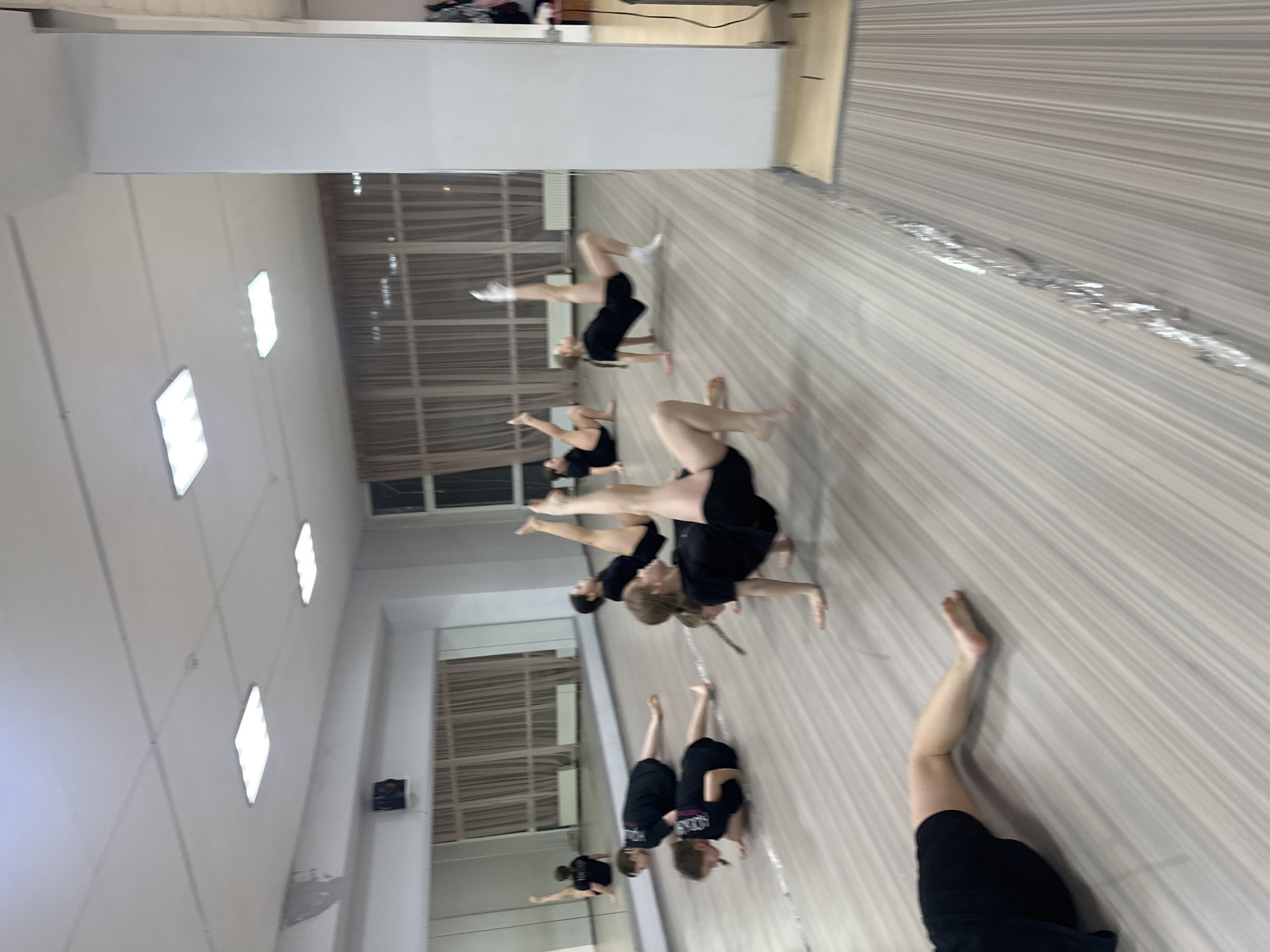 This image depicts an indoor dance or fitness studio located in Votkinsk, Russia. The environment features a light-colored, striped laminate floor with a reflective seam running horizontally in the foreground. A large mirrored wall occupies the left side, reflecting the room and its occupants. White walls are present, with a tall, white structural pillar on the right. Large windows in the background are covered by light beige sheer curtains, revealing a dark exterior, indicating evening or night. The white ceiling contains several square, bright LED light fixtures and a visible smoke detector. A speaker is mounted on the wall above the mirror, and a radiator is positioned below the windows.
Eight individuals, predominantly young females, are present, dressed in black athletic attire (shorts and t-shirts/tops). They are engaged in floor-based exercises or dance conditioning. Multiple individuals are positioned on their hands and feet in an inverted plank or "crab walk" stance, with one leg extended vertically towards the ceiling; some wear white socks, while others are barefoot. Two individuals are in a prone position on the floor in the mid-left, appearing to be in a plank or push-up preparation. One person is partially visible in the immediate foreground, lying on their side with one leg extended. The overall scene suggests a structured physical training session under artificial lighting.