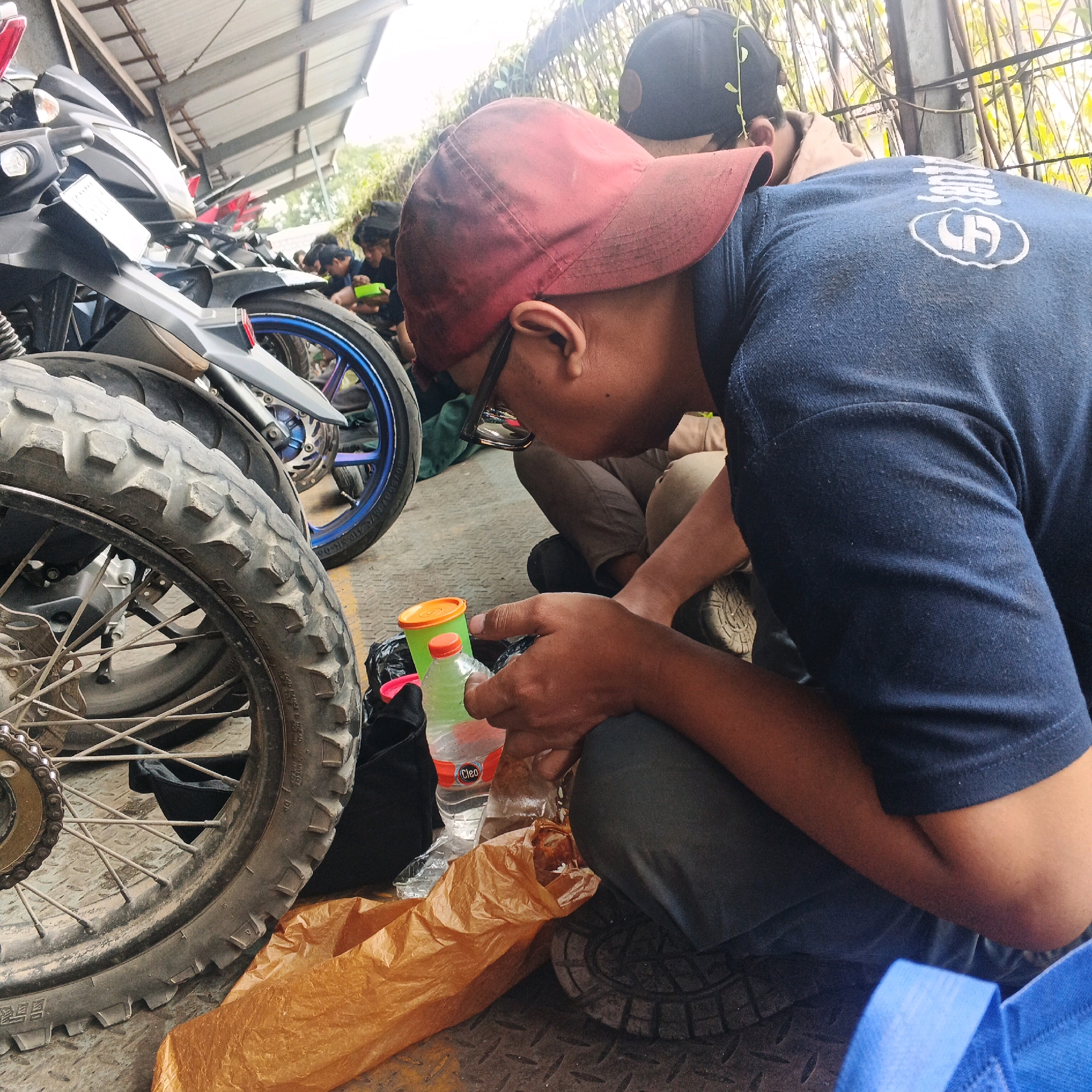 In Banjararum, Indonesia, a close-up shot captures a group of individuals gathered near parked motorcycles under a covered structure. The foreground is dominated by the tire and wheel of a motorcycle, its tread rugged and worn. To the right, a person wearing a red baseball cap and a blue t-shirt with a circular emblem is crouching, holding a brown plastic bag and a clear plastic water bottle with an orange cap. Nearby, another person in a black cap is visible, also crouching. Several other individuals are seen in the background, some appearing to be eating or interacting with each other. The environment suggests an outdoor, possibly utilitarian area, with glimpses of vegetation and metal structures overhead. The lighting is bright, indicating daytime.