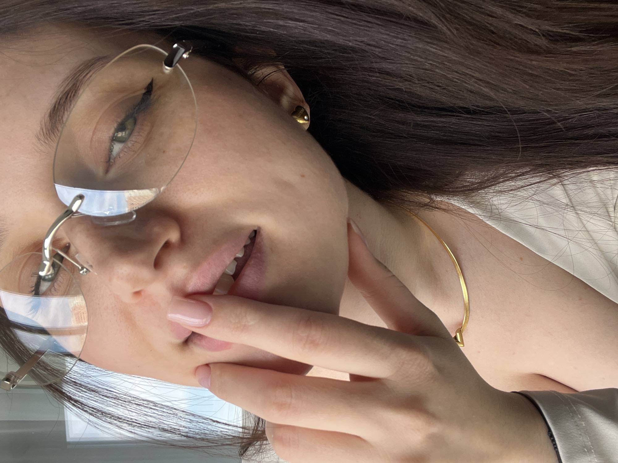 A young woman is the primary subject of the image. She has brown hair, fair skin, and is wearing glasses with a thin silver frame. She also has a gold earring and a gold necklace visible. Her nails are painted a light pink color. Her hand is near her mouth. The reflection in her glasses shows an outdoor scene, possibly buildings and sky. The image is rotated. There is no indication of the city of Tyumen, Russia.