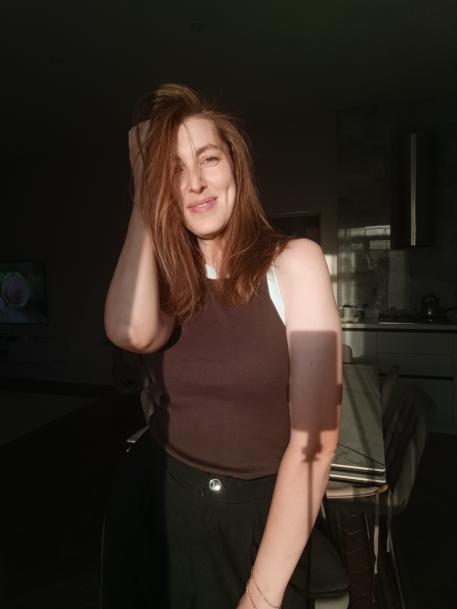 The media file shows a medium shot of a woman with reddish-brown hair, standing in what appears to be an indoor setting, possibly a kitchen or living room. She is looking directly at the camera with a slight smile, her left hand is raised and touching her hair. The lighting suggests it is late afternoon or early evening, with strong, directional sunlight casting shadows across the scene.
The woman is wearing a dark brown ribbed tank top with a light blue strap visible at the neckline, and dark pants. A glint of light is visible on a button on her pants. She is wearing a delicate bracelet on her left wrist.
The background is dimly lit, with a kitchen counter and cabinets visible on the right side, suggesting a domestic environment. A stainless steel hood is above the stove area. On the left side of the image, a television screen is visible, displaying an image of a halved walnut.
The overall mood of the image is relaxed and contemplative, enhanced by the warm glow of the sunlight. The presence of indoor elements like the television and kitchen appliances anchors the scene in a home setting. No visible text is present in the image.