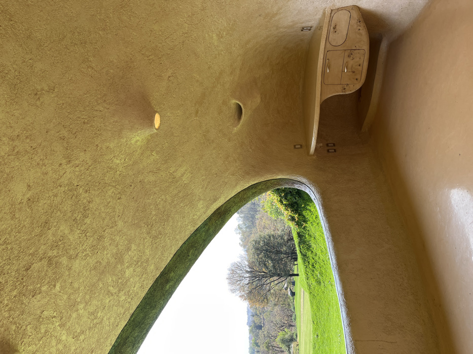 The image depicts an interior view within the Casa Orgánica (Organic House) by Javier Senosiain, located in Naucalpan de Juárez, Mexico. The scene is characterized by biomorphic, curvilinear architecture, where walls, ceiling, and floor flow continuously in a single, untextured, light tan or beige finish, suggesting a monolithic construction of plaster or concrete.
On the left, a large, irregularly shaped opening acts as a window, framing an exterior landscape. The exterior displays a vibrant green, undulating terrain, likely a manicured lawn or golf course, transitioning into a tree-filled background. Several deciduous trees with sparse foliage and evergreens are visible under a bright, overcast sky.
The right side of the interior features a recessed, warm-toned circular light fixture in the ceiling, along with a deeper, smaller circular indentation below it. A built-in, light-colored desk or counter, with an organic, wavy upper surface, extends from the wall. Below the main desk surface, a light-toned wooden cabinet is integrated, featuring two small drawers with silver pull handles on the left and a larger, irregularly shaped door or drawer with a silver pull handle on the right. Several standard electrical outlets or switches are mounted on the wall, both above and below the desk structure. The floor is a continuous, smooth surface matching the walls and ceiling, reflecting ambient light. No individuals or loose objects are present in the scene.