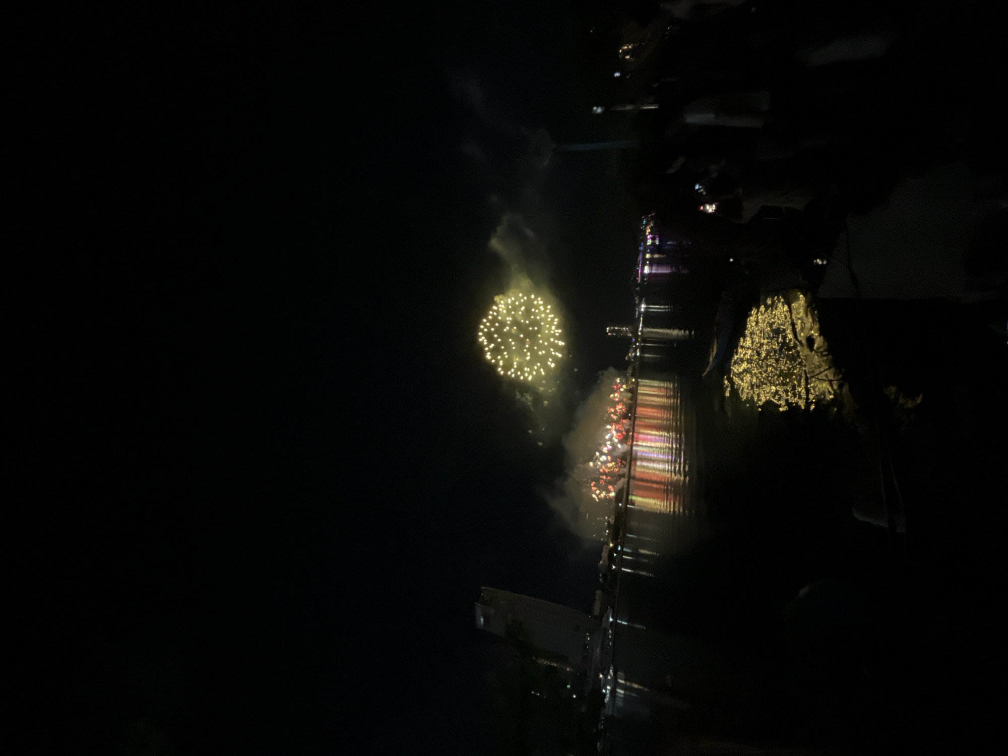 This image captures a nighttime fireworks display over a body of water, identifiable as the Han River in Đà Nẵng, Vietnam.
In the upper-middle of the frame, a large, spherical fireworks burst illuminates the sky with numerous small, bright yellow-gold lights, surrounded by a cloud of smoke. To the left, closer to the water, a series of smaller, predominantly red and white fireworks bursts are visible, also emitting significant smoke. The vibrant lights of the fireworks reflect strongly on the dark surface of the Han River, creating elongated streaks of yellow, red, and white.
On the left bank of the river, the silhouette of a tall, modern building, characteristic of the Novotel Danang Premier Han River hotel, stands against the dark sky. Along the opposite bank, numerous city lights and illuminated structures, including what appears to be a bridge in the distance, create a glowing backdrop.
The immediate foreground on the right side is dark, but the silhouettes of several spectators are discernible. At least one individual is visibly holding up a smartphone, likely capturing the event. A smaller boat or kayak is subtly visible on the river's surface in the mid-ground, reflecting the golden light from the fireworks. The overall scene is one of a festive night event with pyrotechnics illuminating the city riverfront.