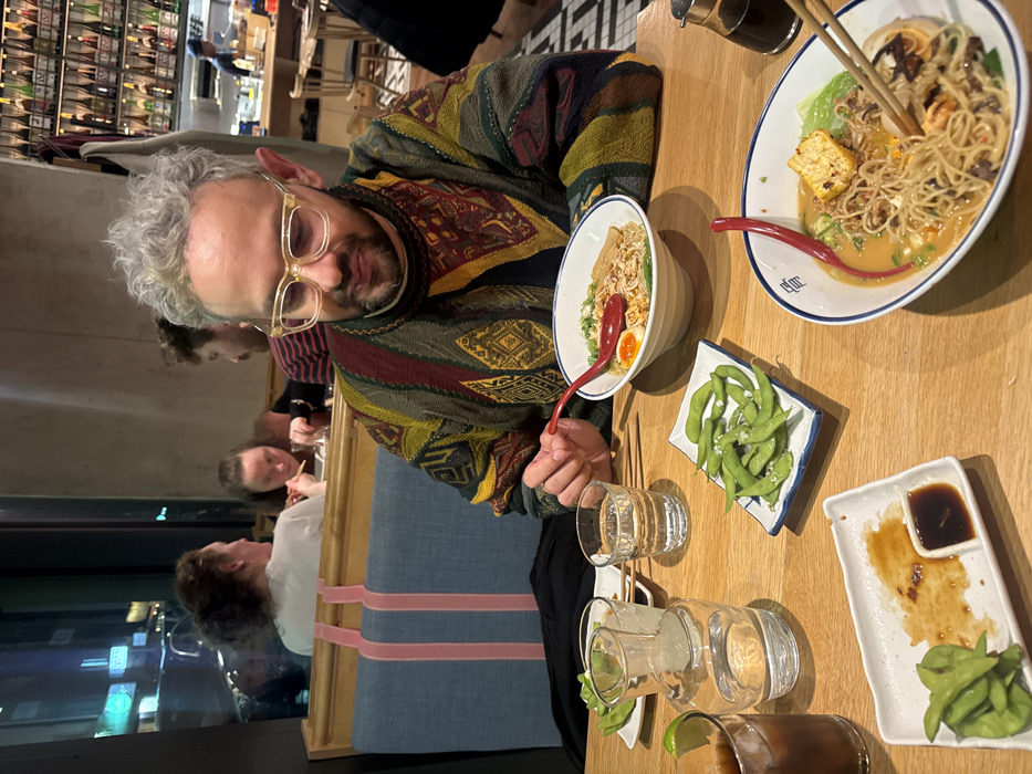 A person wearing glasses and a patterned sweater sits at a restaurant table. Two bowls of ramen are visible, along with a small dish of edamame. Several drinks sit on the table. Other people are seated at the table in the background. The restaurant appears to be located in an urban area at night.