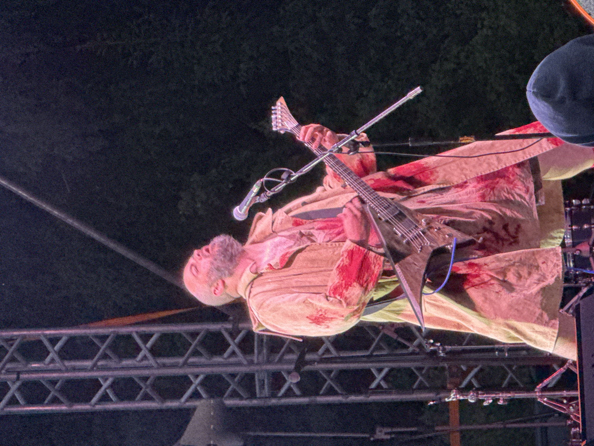 A male performer with a bald head and a full beard is captured mid-performance on an outdoor stage in Большая Кивара, Russia. He is wearing a long, light-colored (tan or beige) coat or duster heavily splattered with red simulated blood, over what appears to be a similarly blood-splattered red/pink shirt. The performer's head is tilted back, mouth open, suggesting he is singing or screaming into a black microphone positioned on a multi-armed stand directly in front of him. He is playing a distinctive V-shaped electric guitar with a pointed headstock and multiple strings (likely 7 or 8-string), connected by a visible blue cable. His right hand is on the fretboard, and his left hand is near the guitar's body.
To the left of the performer, a tall, grey metal truss structure, typical of concert staging, is visible. The background is dark, indicating night-time, with indistinct dark foliage or trees visible to the right. Various pieces of stage equipment, including parts of a drum kit (stand, drum shell) and other stands, are partially visible behind and around the performer. In the lower right foreground, the top of a person's head, covered by a dark blue hooded garment or beanie, indicates an audience member. The scene depicts an intense live musical performance, likely a rock or metal concert, characterized by theatrical elements such as the "blood" splatters on the costume.