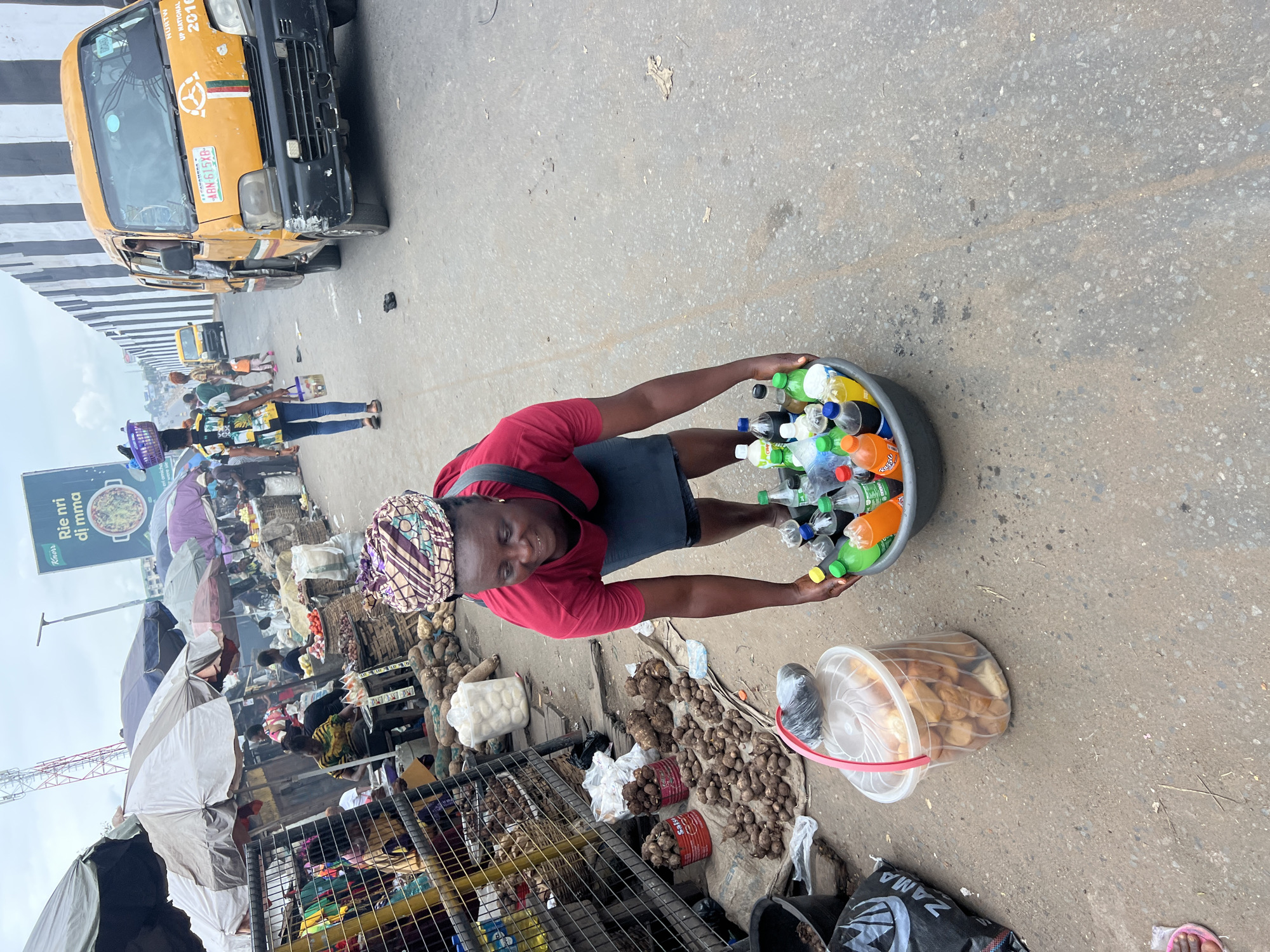 A woman in a red shirt and head wrap holds a basin filled with bottles of various colored drinks. She is standing on a street in Nkpor, Nigeria. There are other vendors in the background along with vehicles and pedestrian traffic. A yellow bus is visible in the upper left. Beside the woman is a container filled with what appears to be fried food. The street is paved, and there are other objects scattered on the ground.