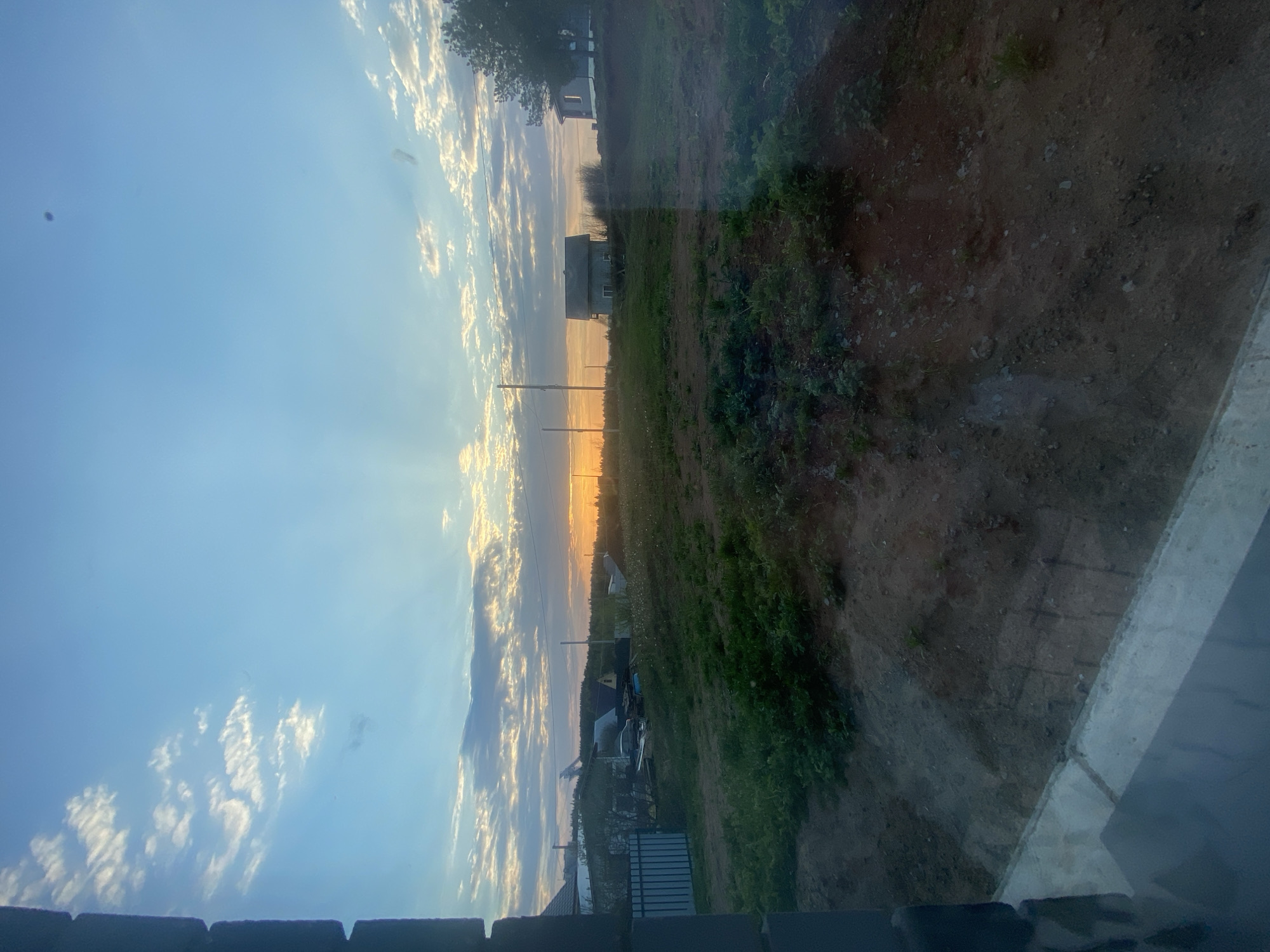 The image presents a view from inside a building, looking out onto a landscape at sunset or sunrise. The sky occupies the left side of the image, showing a gradient of blue transitioning into orange and yellow hues along the horizon, with scattered clouds. The land area consists of a grassy field with some vegetation, transitioning into a dirt area on the right side. Houses and infrastructure are visible in the distance near the horizon line. There are power lines and a fence. The location is unknown, and there are no people visible.