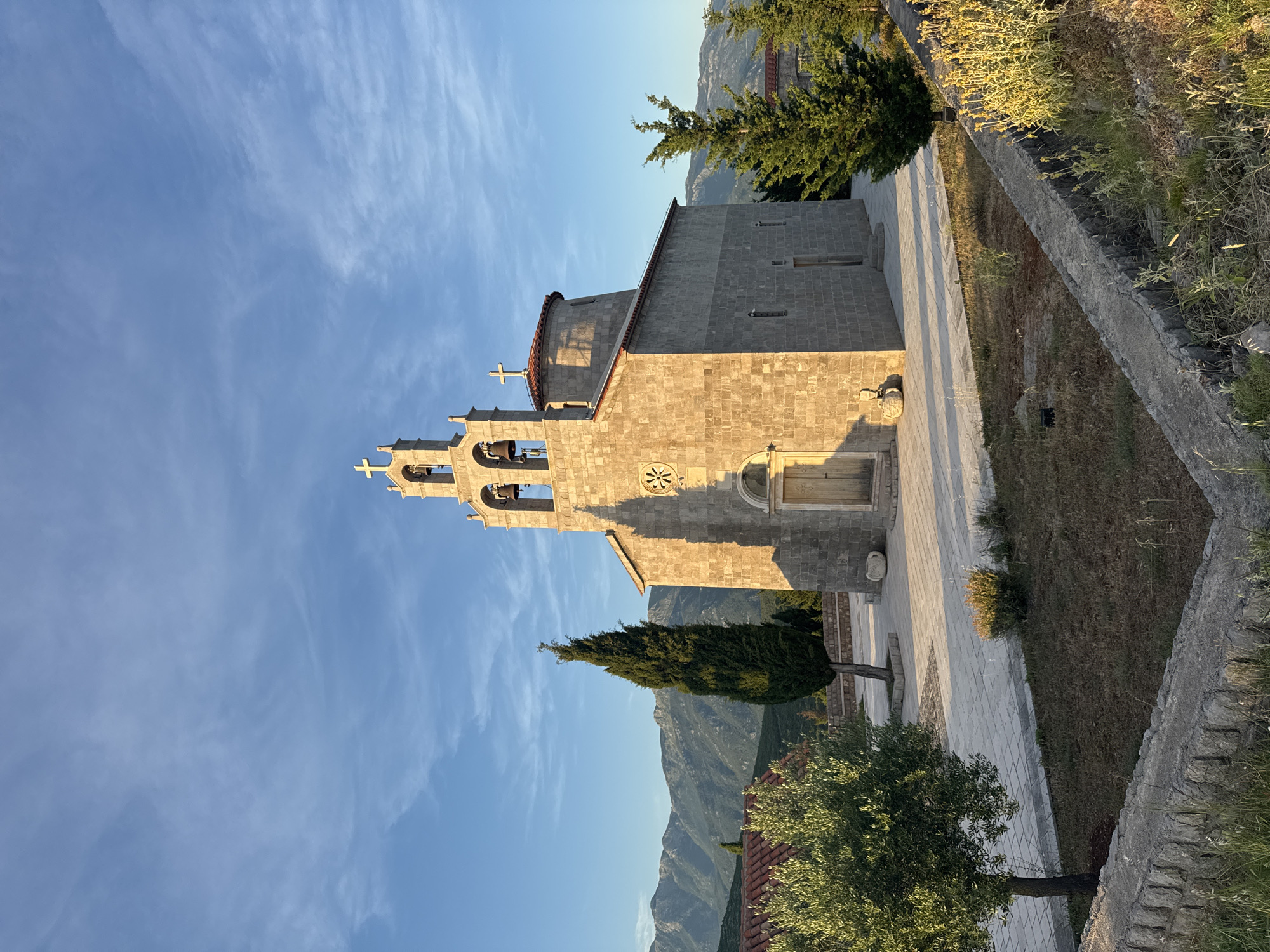 The photograph captures a stone church with a bell tower situated in Kovačko Polje, Montenegro. The building is constructed from light-colored stone blocks. The bell tower, topped with a cross, features three bells. Adjacent to the main structure is a smaller domed section, also crowned with a cross. The church has a wooden door and a circular decorative element above it. The surroundings include a paved courtyard, a cypress tree, an olive tree, and other greenery. In the background, a range of mountains stretches across the horizon under a blue sky with scattered clouds. The scene is illuminated by the warm light of what appears to be late afternoon or early morning. A low stone wall runs across the foreground.