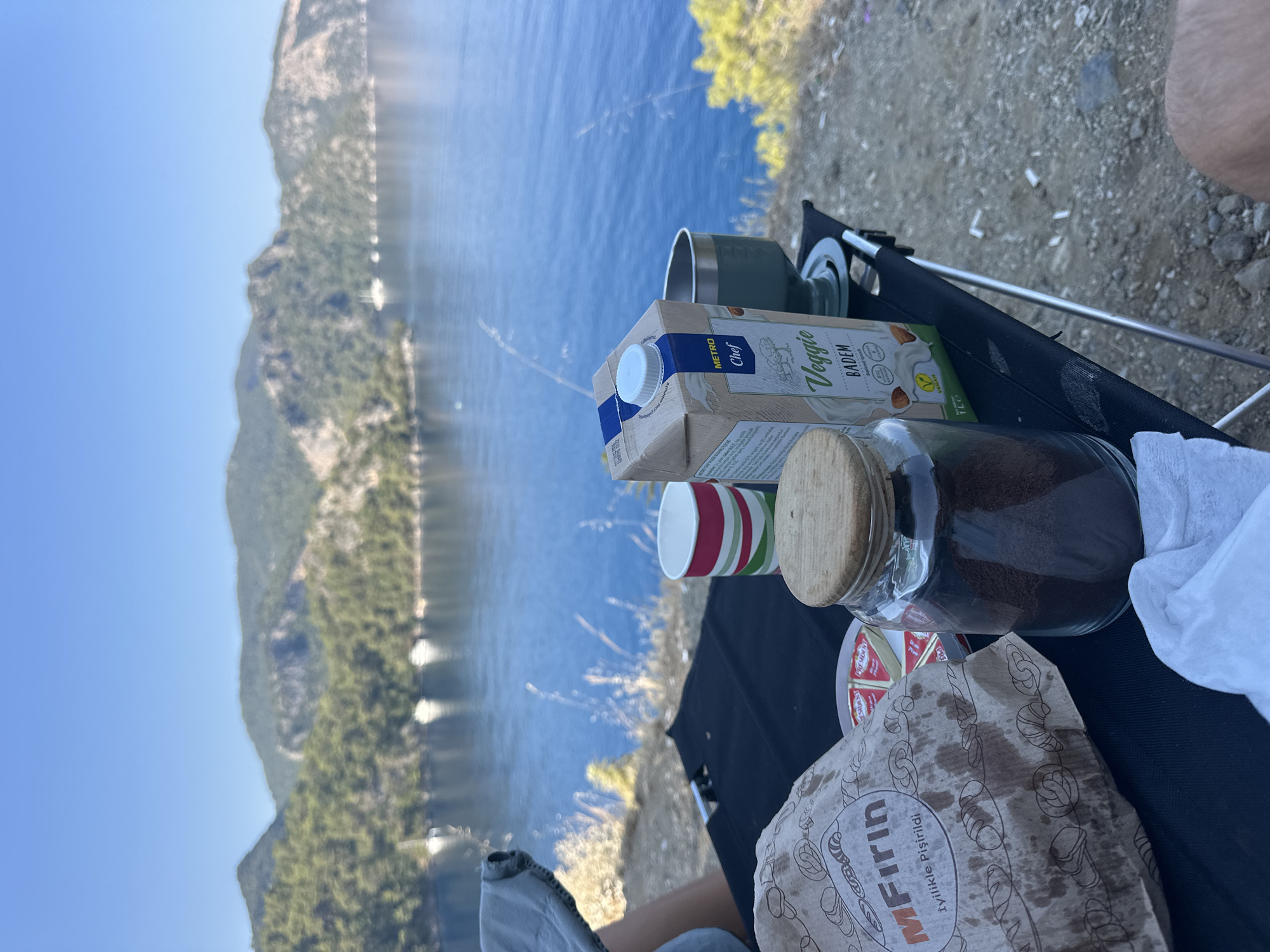 A picnic is set up on a folding black table at an elevated viewpoint overlooking a body of water with a mountainous, tree-covered coastline. In the foreground, on the table, are a brown paper bag with the logo "MFırın" which translates to "My Oven" and the text "İyilikle Pişirildi" meaning "Cooked with Goodness", a small plate with several triangular wedges of cheese, a clear glass jar filled with dark brown granular material, a red, white, and green striped paper cup, a carton of "METRO Chef Veggie BADEM" almond milk, and a green metal camping mug. A white napkin is also visible on the table. The background shows the blue water with several small white boats sailing on it, and the verdant, hilly landscape of the Fethiye coastline under a clear blue sky. The scene suggests a peaceful outdoor meal in a scenic location.