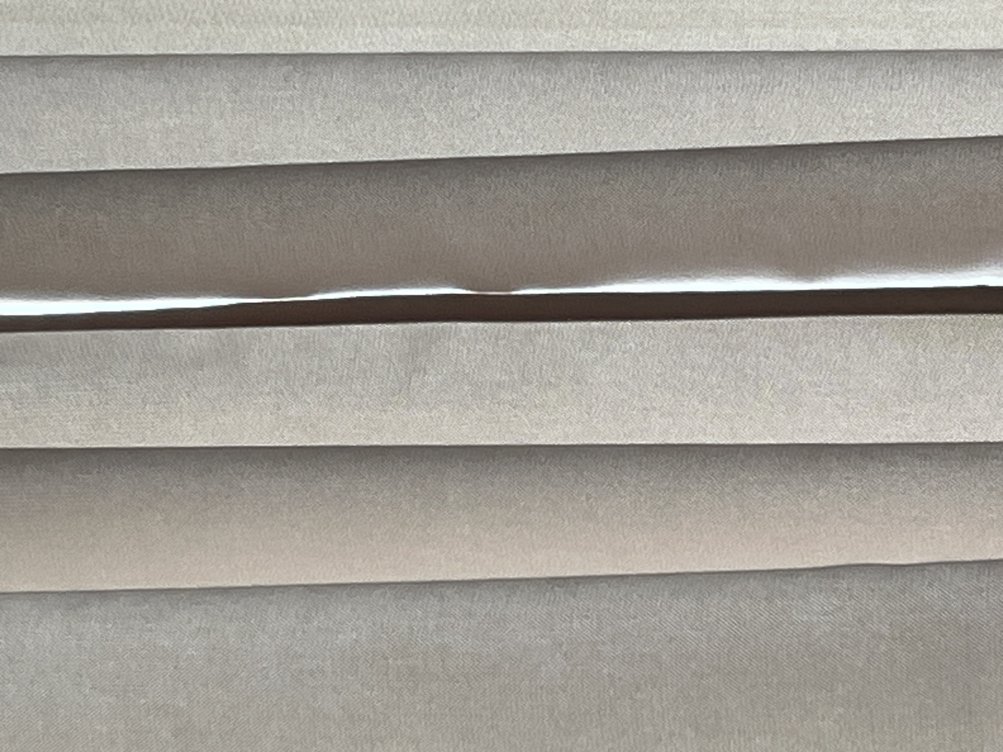 The image is a close-up, full-frame shot of vertical blinds. The blinds are made of a textured fabric in a neutral, light brown or beige color. There are multiple panels of the blinds visible, with slight variations in shadow and light due to their texture and the way they are positioned. A thin sliver of bright light is visible between two of the blinds, suggesting a light source behind them. The texture of the fabric appears somewhat coarse, with a subtle weave pattern visible upon close inspection. The composition is abstract, focusing on the repetitive vertical lines and the play of light and shadow.